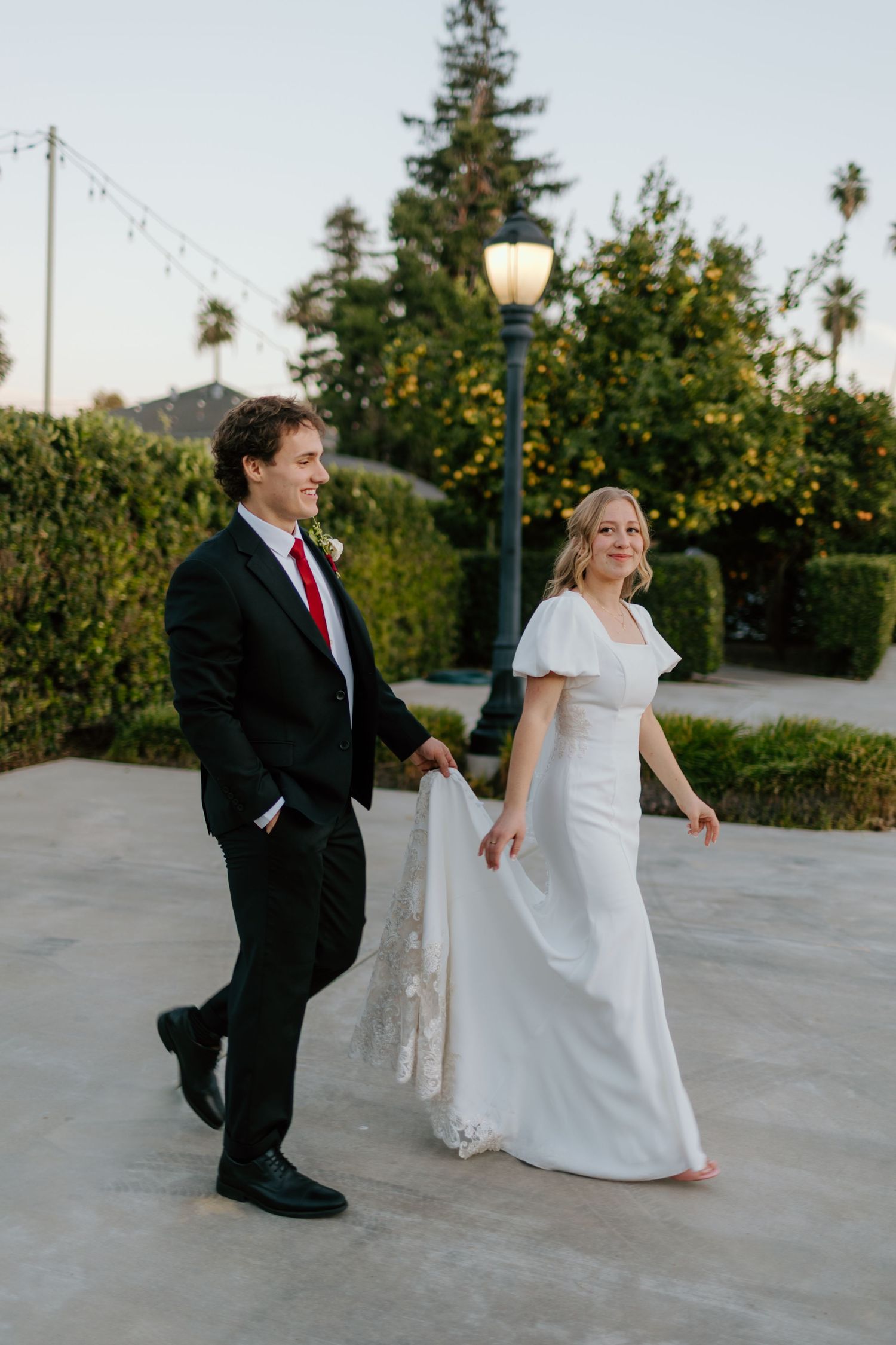 Bride in white gown and groom in black suit holding hands walking outdoors at golden hour wedding.
