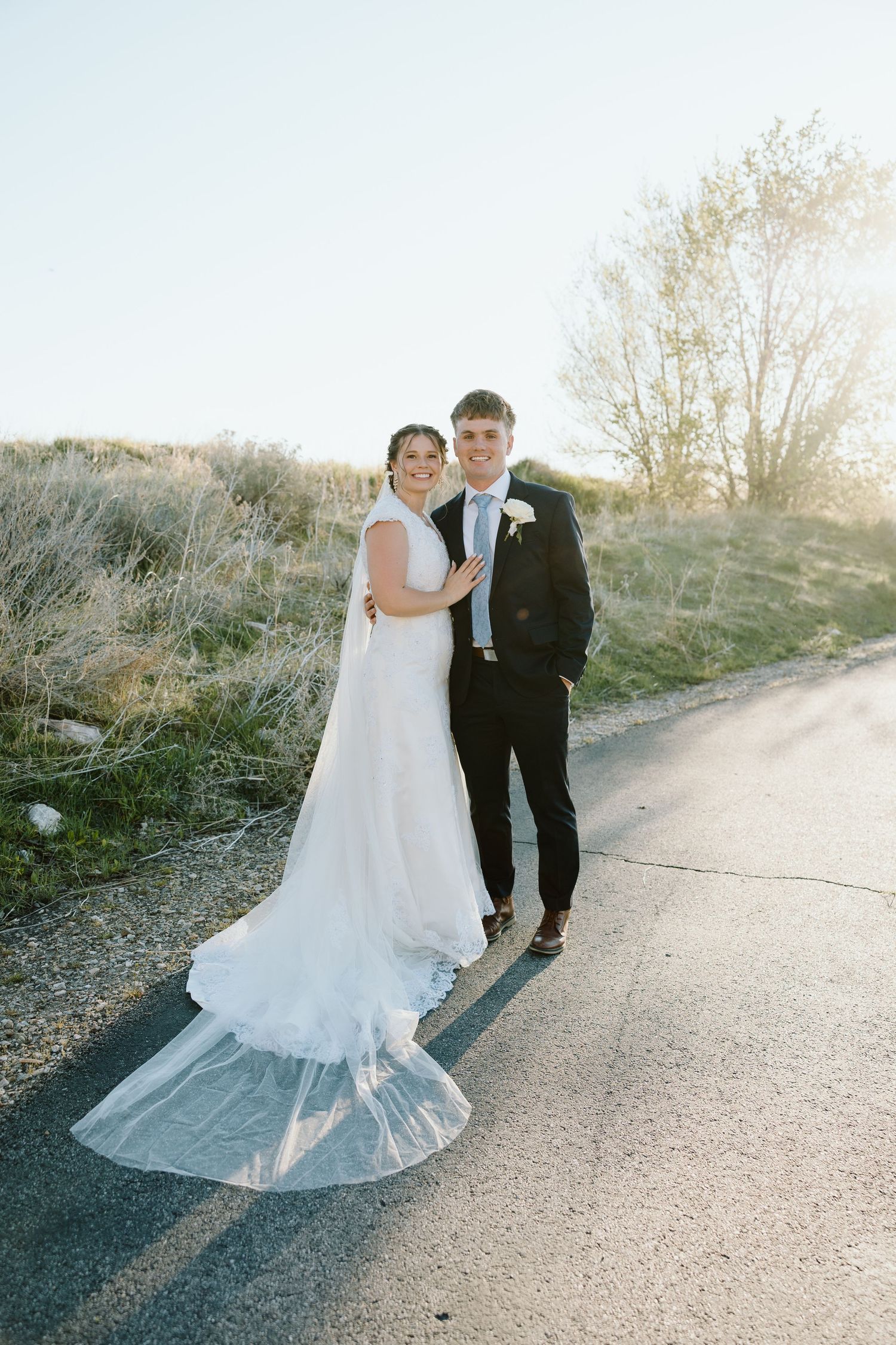Bride and groom pose together on a sunny road, bride wearing long veil and white gown.
