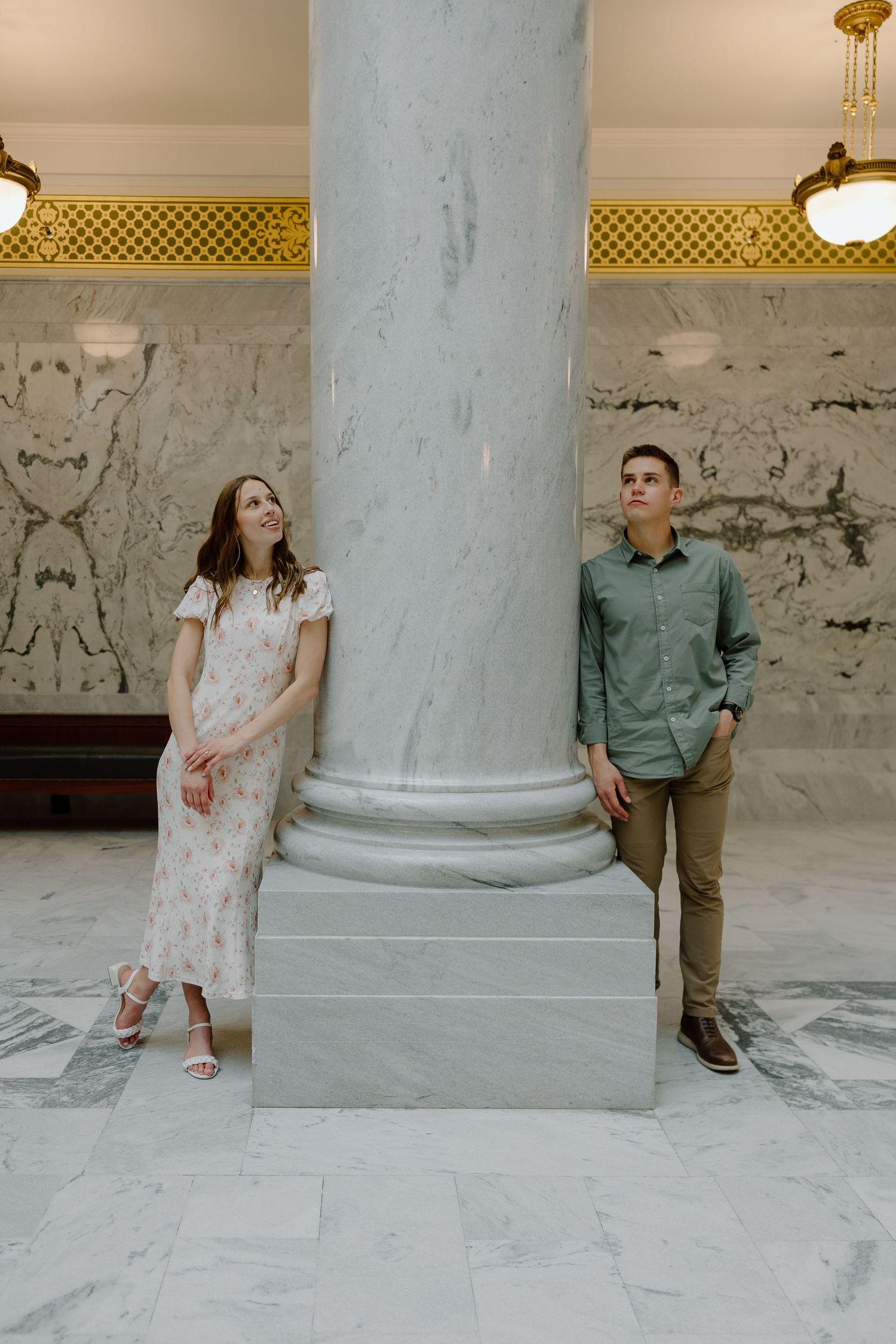 Couple standing on opposite sides of a large marble column in a classical building interior.
