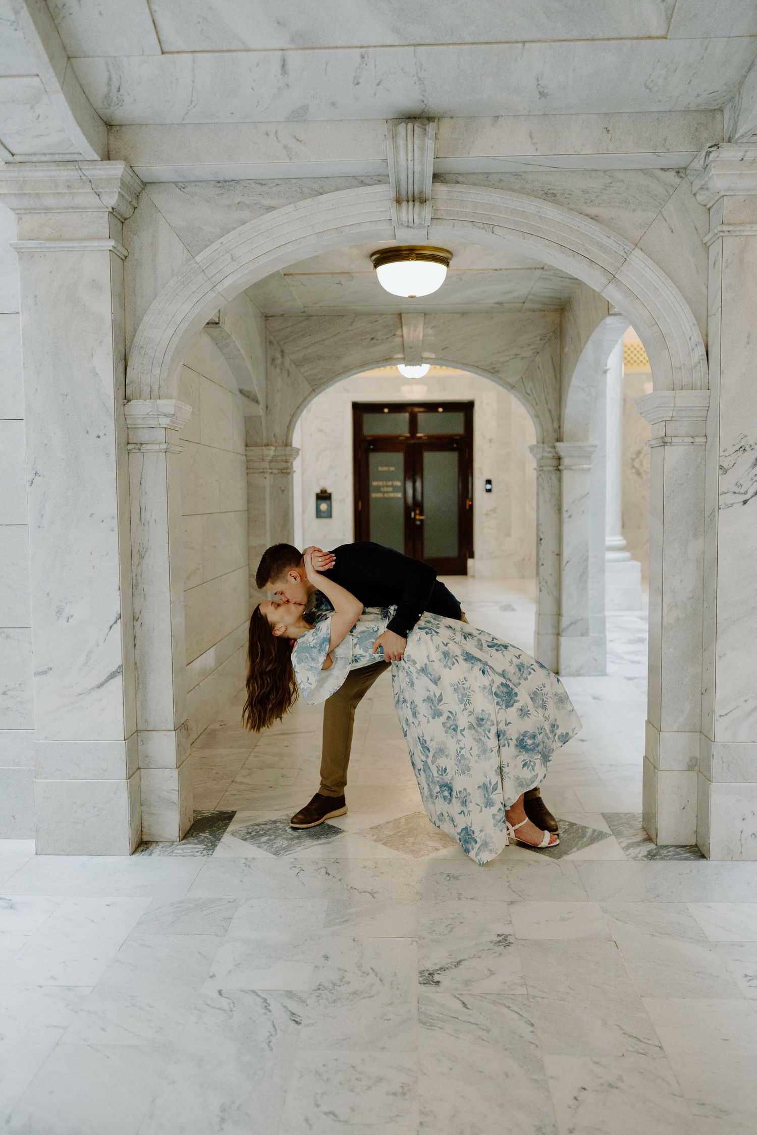 A man dips a woman for a romantic kiss inside an elegant marble archway corridor.