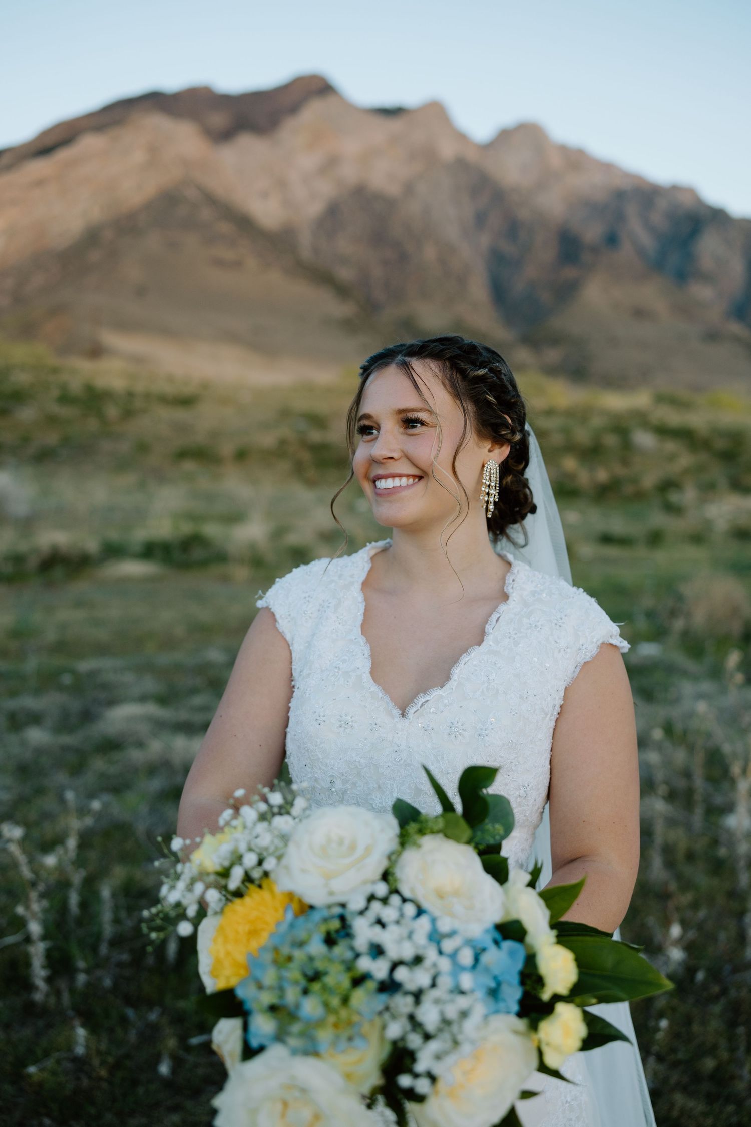 Smiling bride in lace wedding dress holding blue and white bouquet with mountain backdrop.