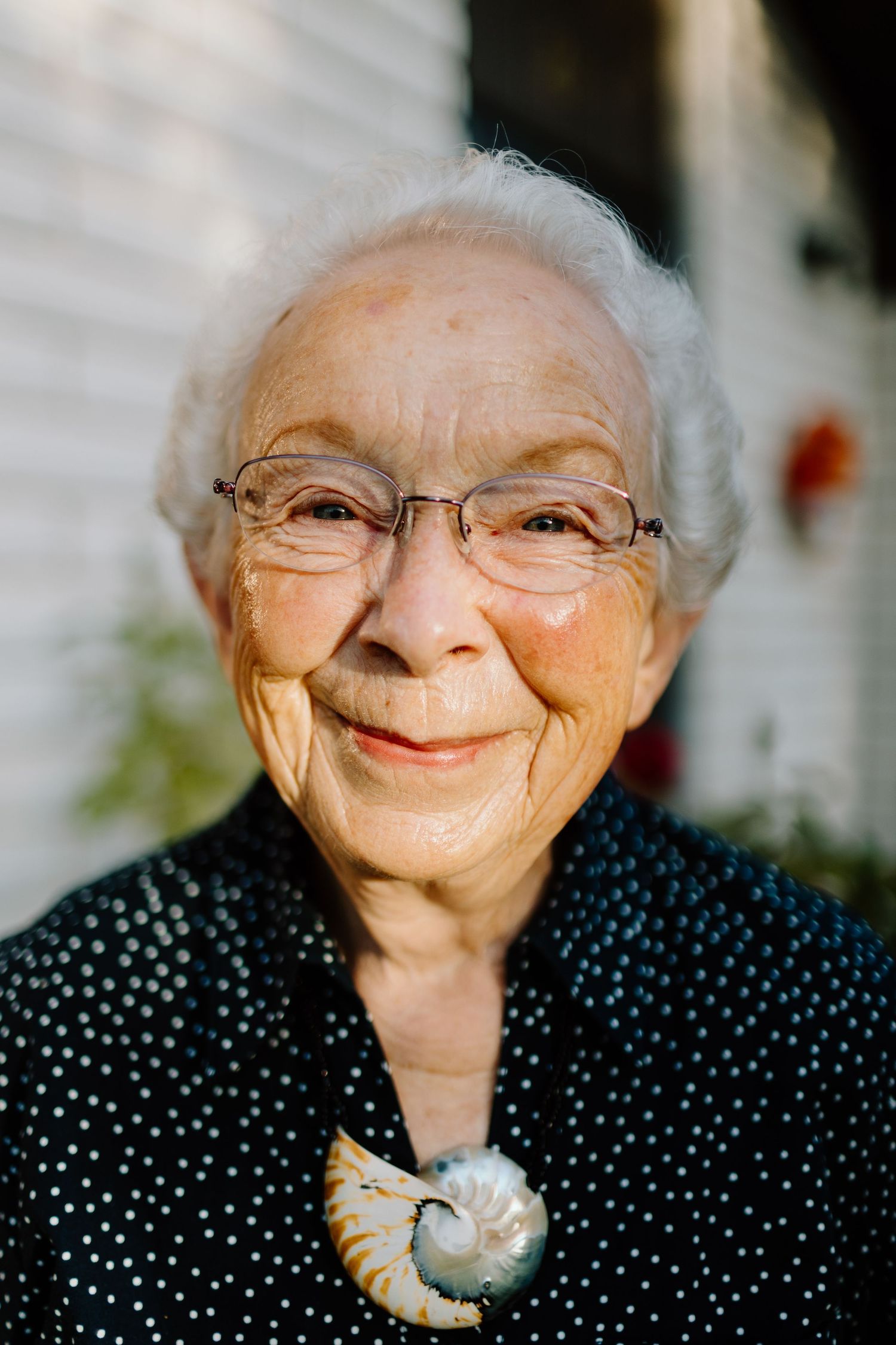 An elderly woman in a polka dot blouse smiles warmly at the camera.