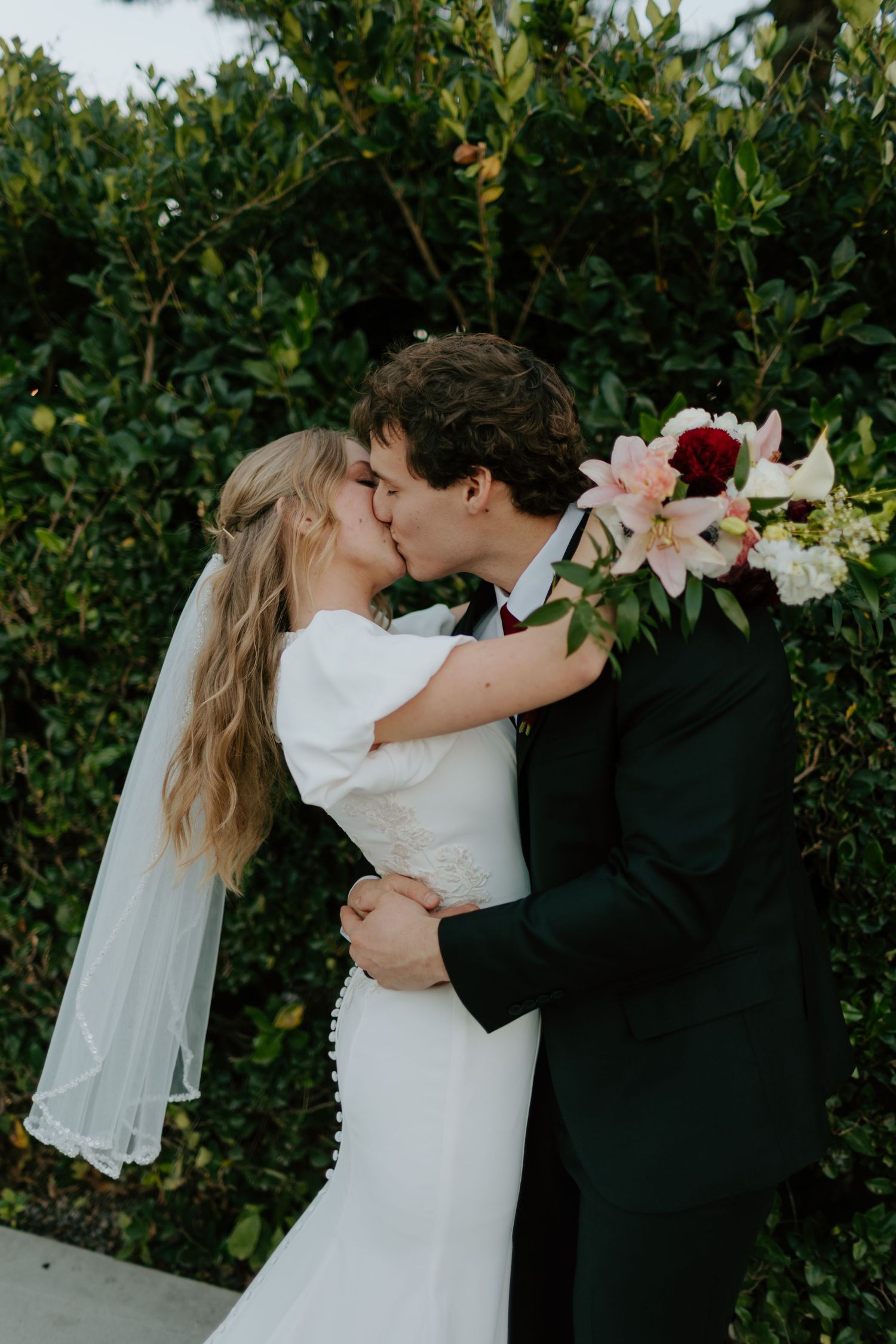 Bride and groom kissing in front of lush green hedge, bride holding pink lily and white floral bouquet.