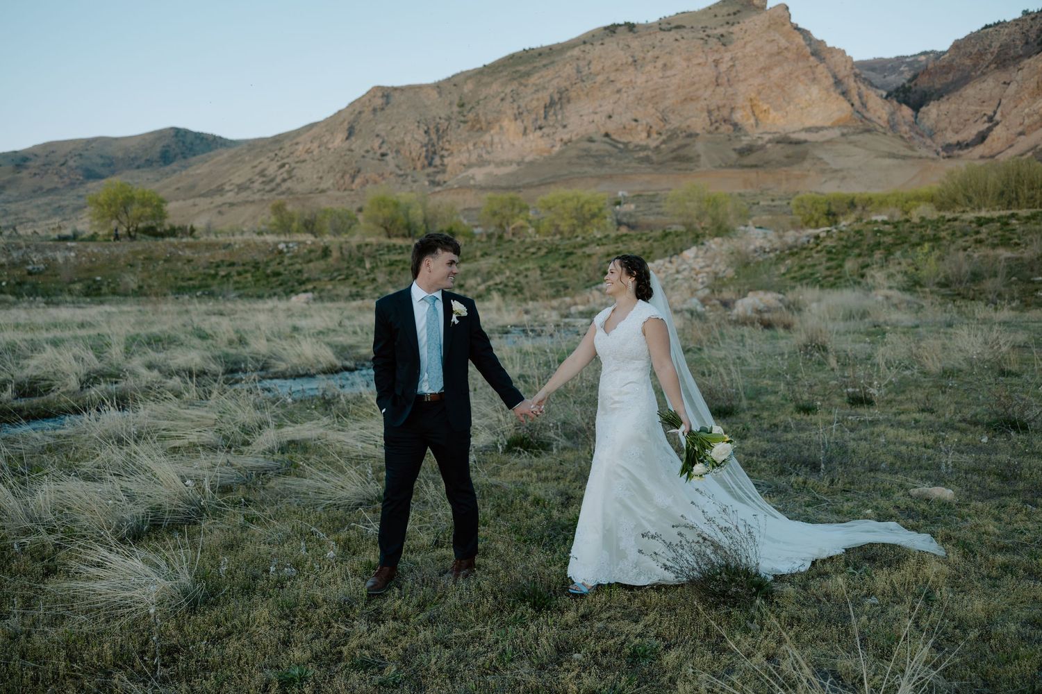 Bride and groom hold hands in a scenic desert landscape with rocky mountains, wearing wedding dress and black suit.