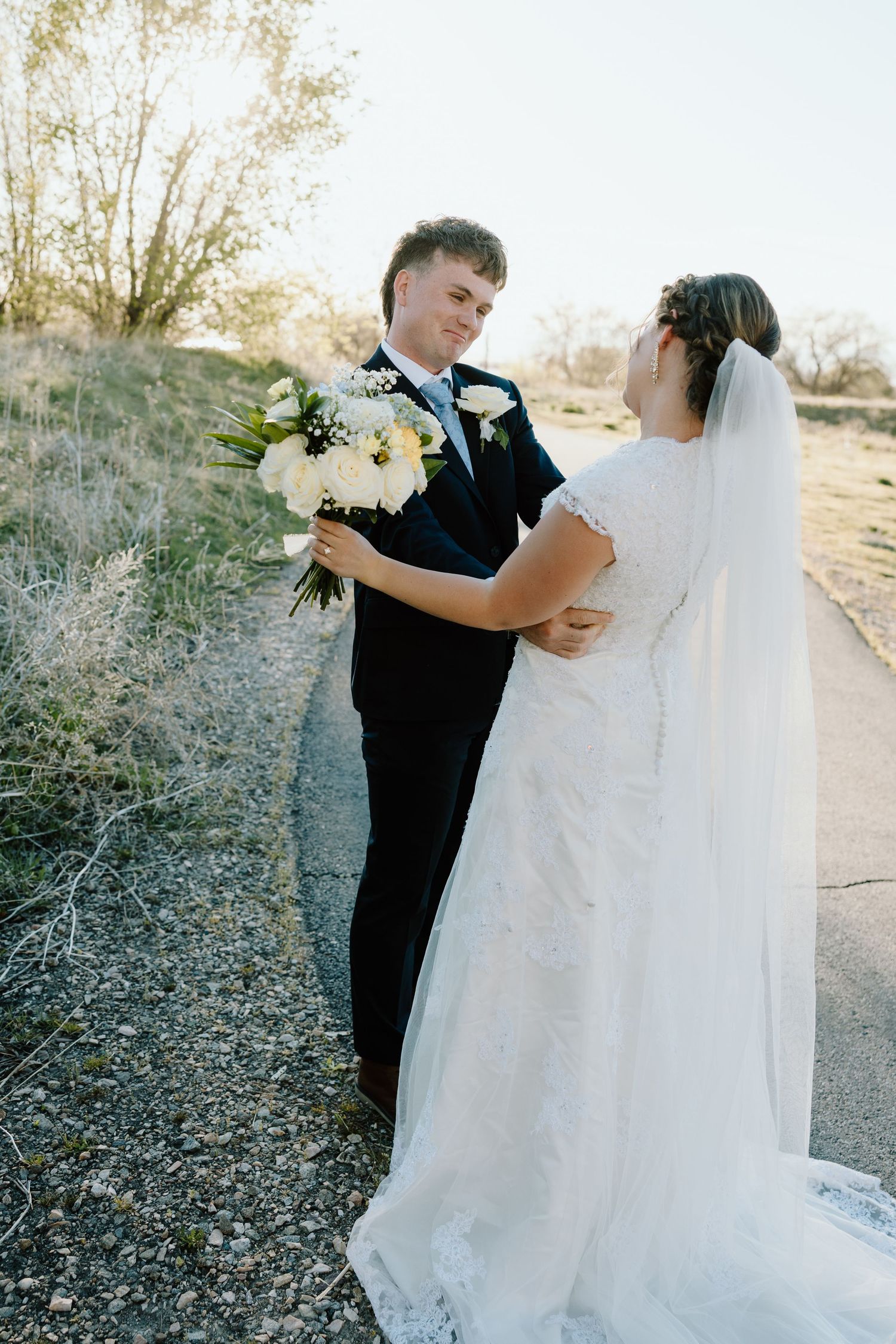 Groom in black suit hands white bouquet to bride in lace wedding dress on outdoor path.