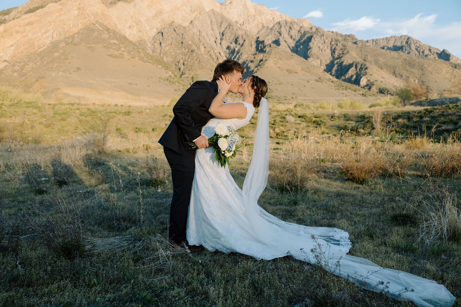 Groom dipping bride in white gown for a kiss in golden field with scenic mountain backdrop at sunset.