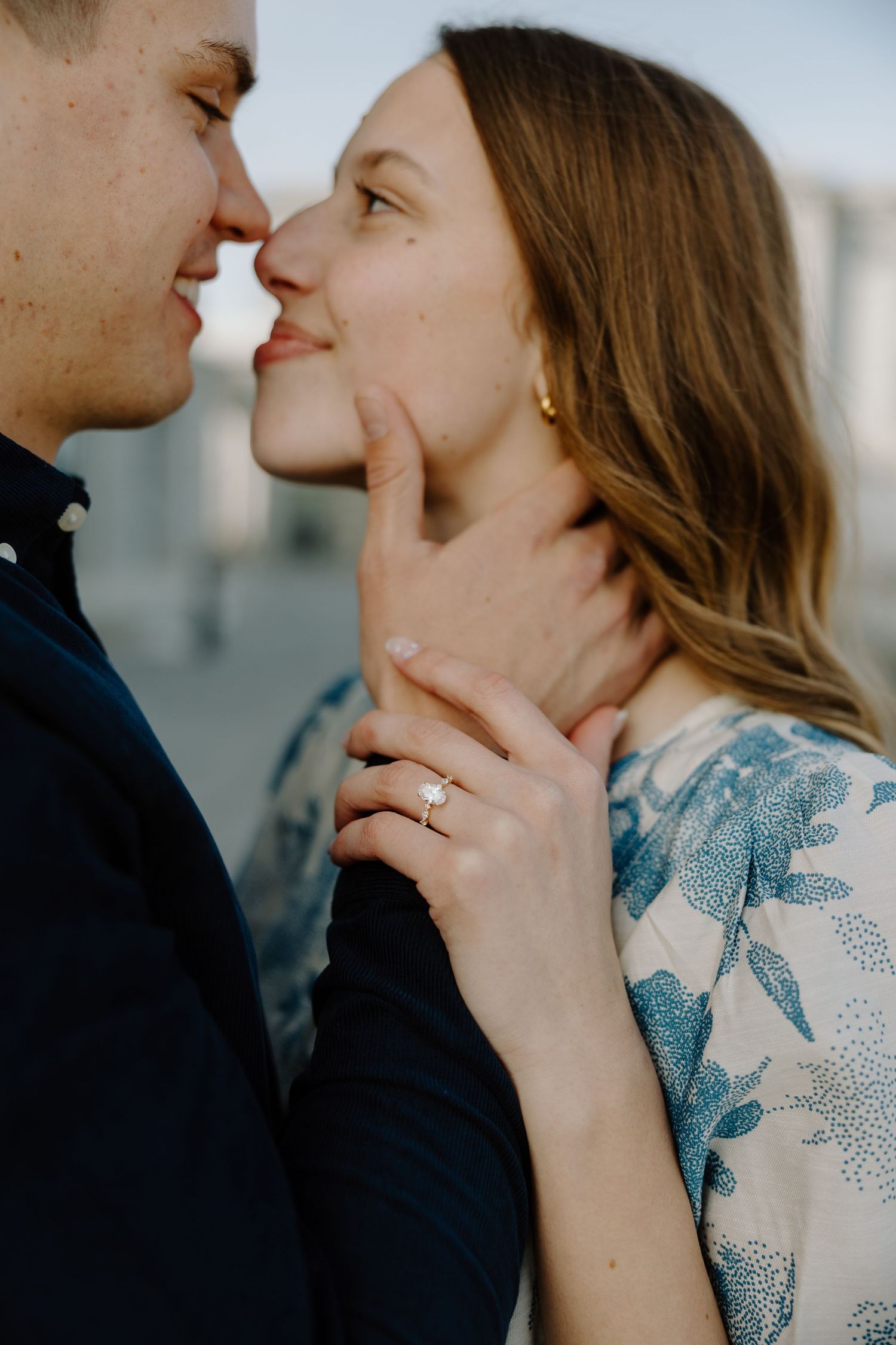 Close-up of couple nearly kissing, woman wearing engagement ring in blue floral dress, romantic moment.