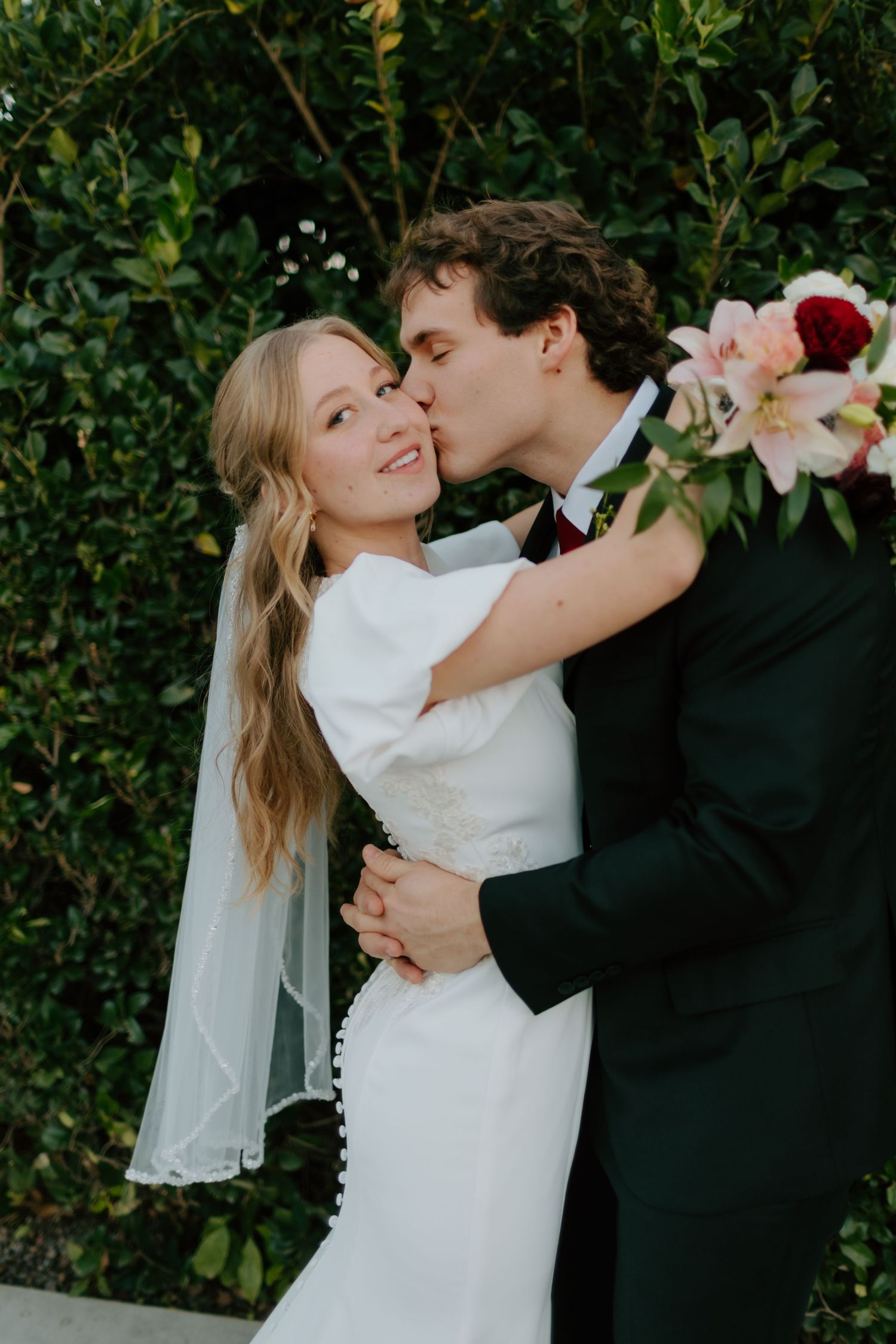 Groom in black suit kisses bride on cheek as she smiles, standing before lush green hedge with floral bouquet.