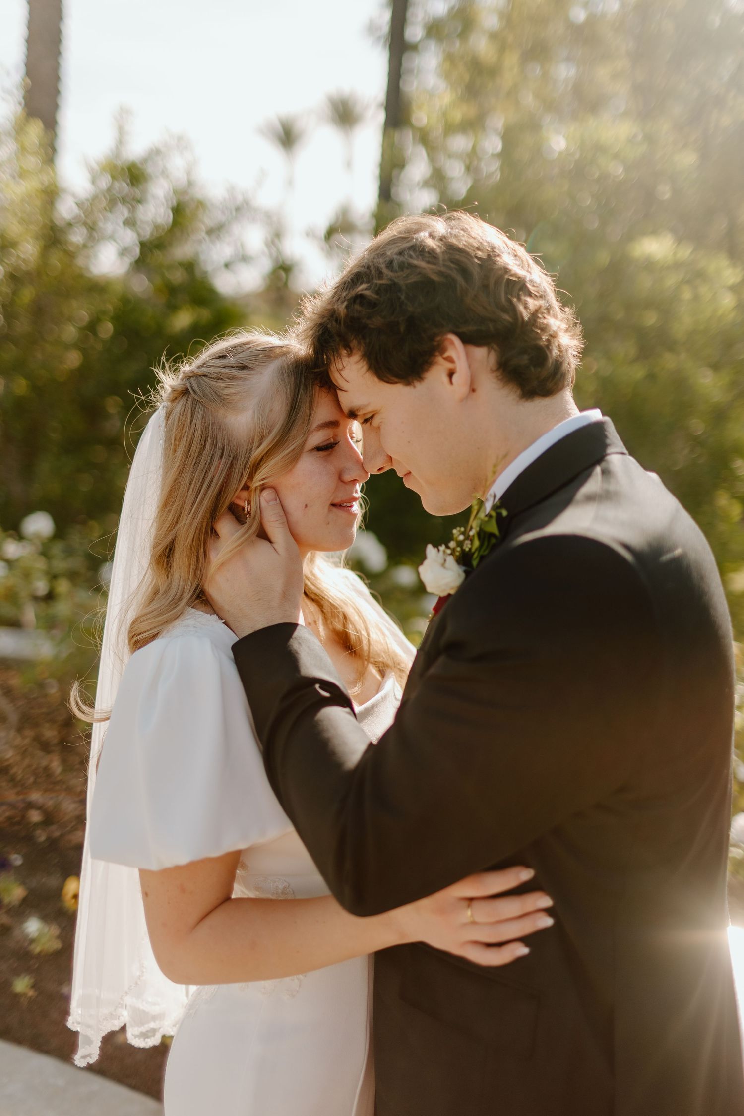 A bride and groom share an intimate moment, touching foreheads in golden sunlight, surrounded by lush greenery.