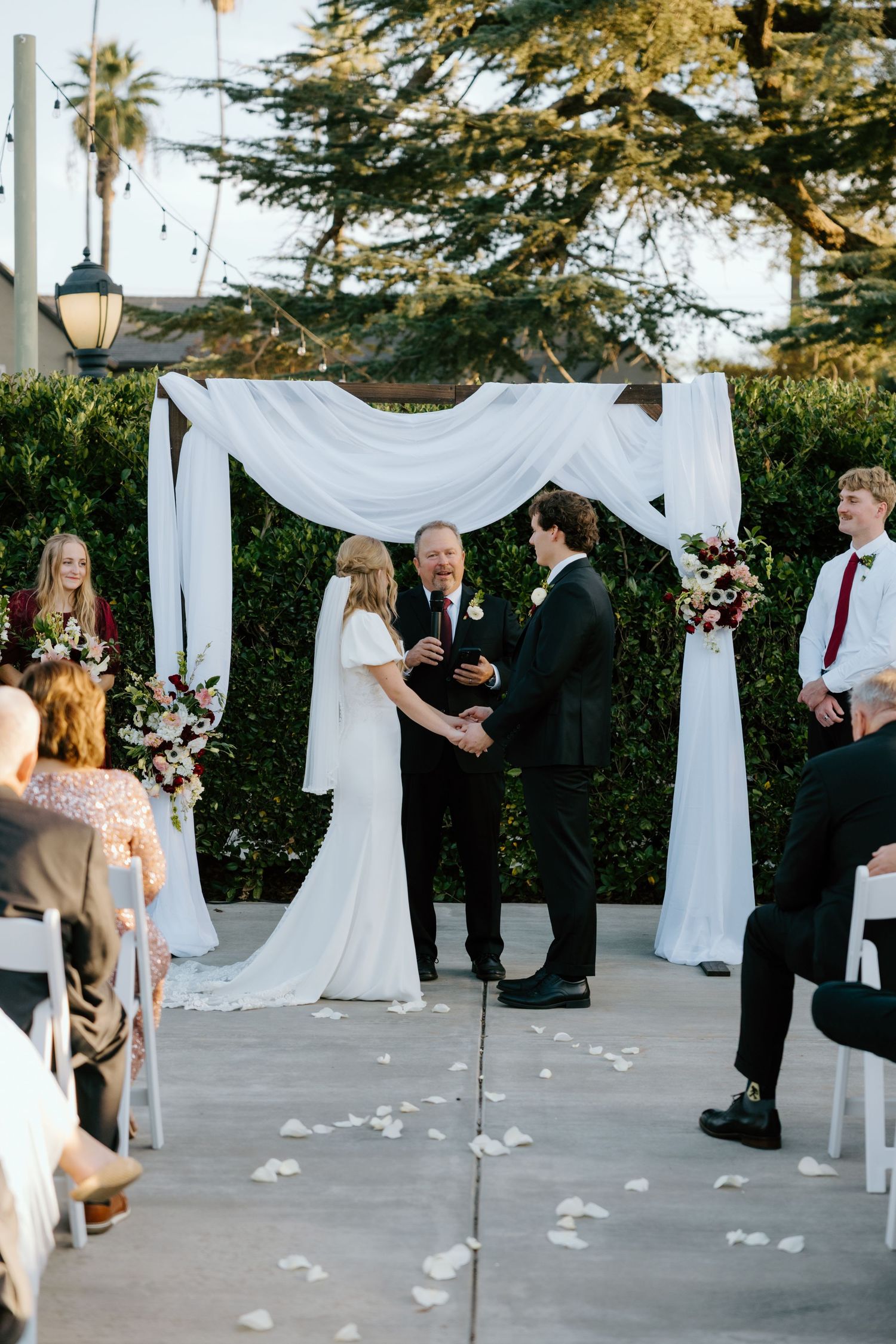 Bride and groom exchange vows under a white draped arch surrounded by wedding party at outdoor ceremony.
