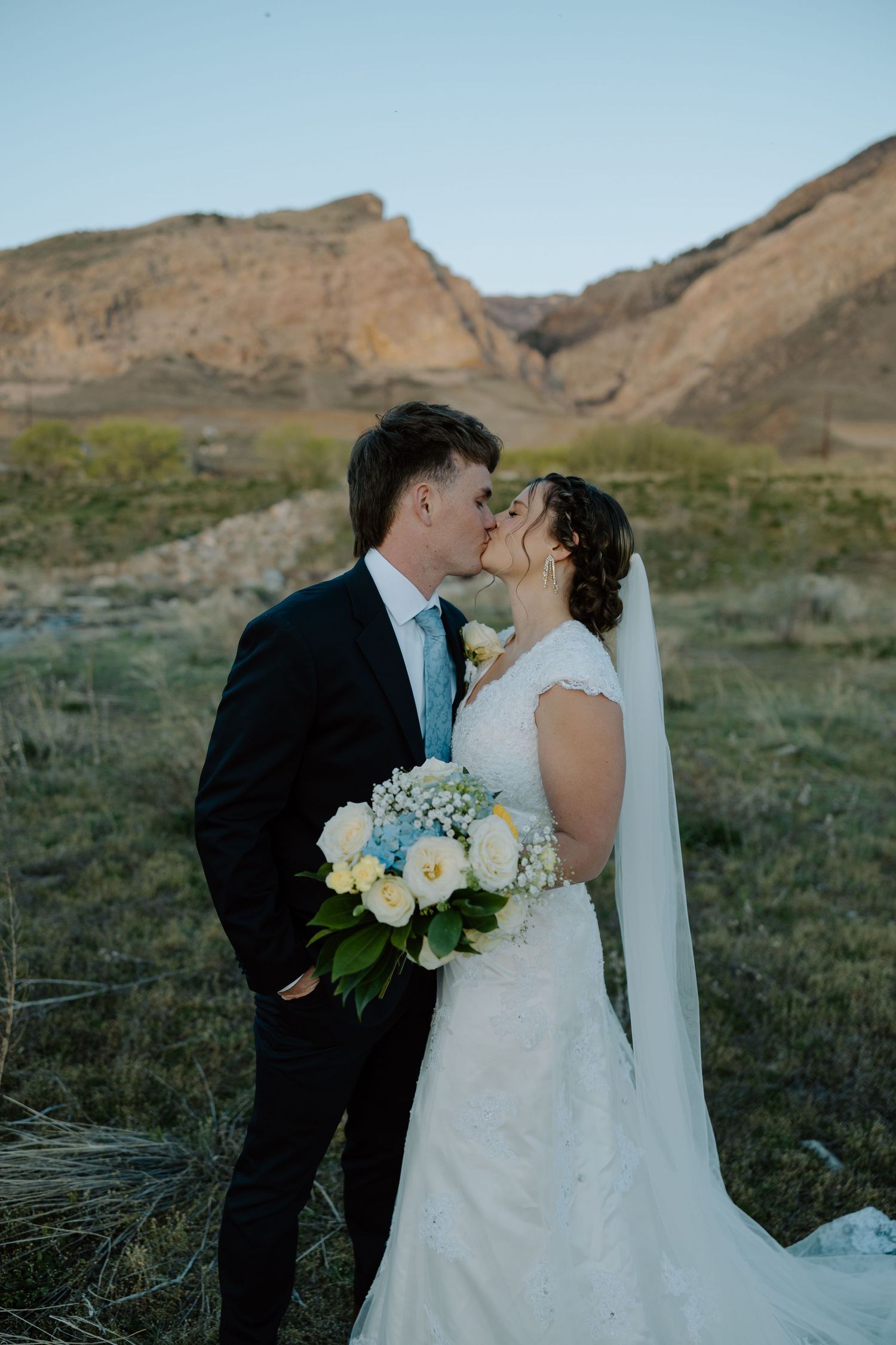 Newlyweds share a romantic kiss holding a white and blue bouquet with rocky mountains behind them.