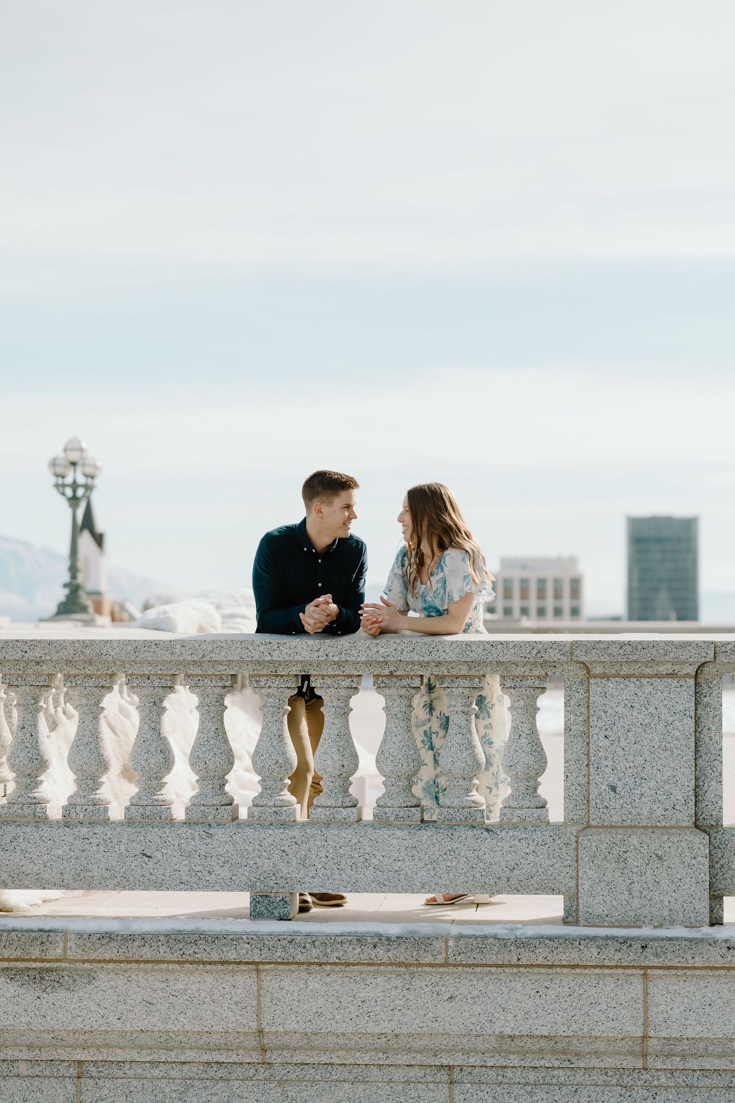 Couple leaning on stone balustrade balcony talking and holding hands at a grand building outdoors.