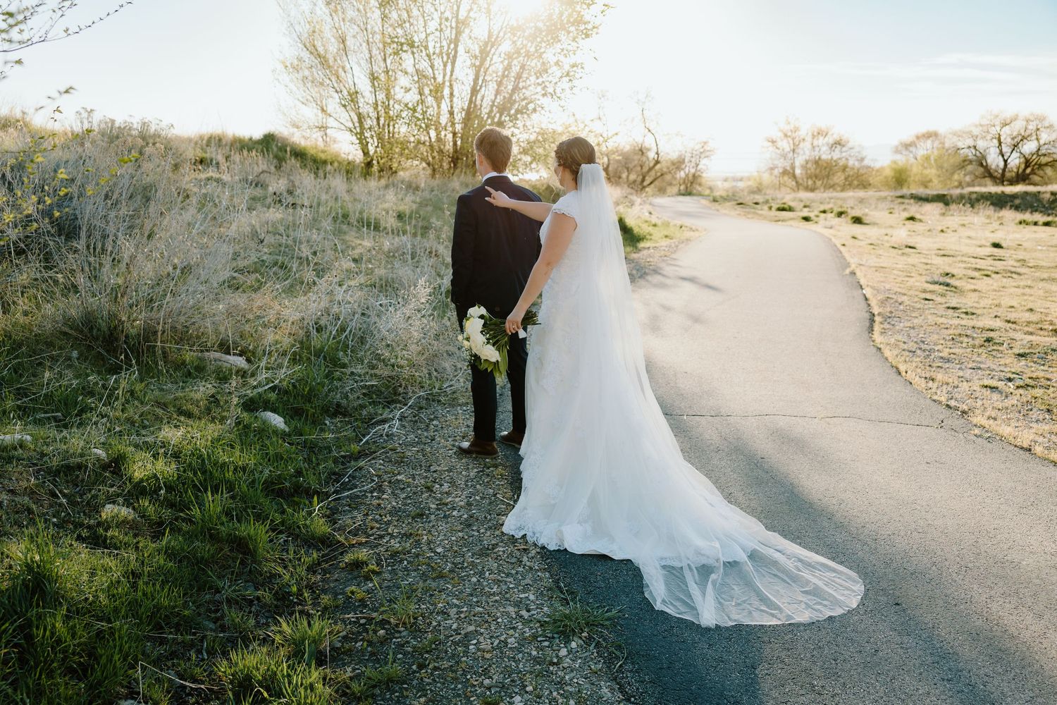 Bride and groom walking hand in hand down a scenic path, bride's white gown trailing behind them at golden hour.