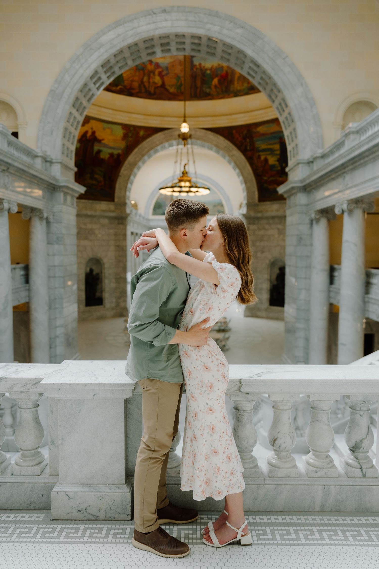 Couple sharing a romantic kiss inside a grand marble building with ornate arched ceilings and chandelier.