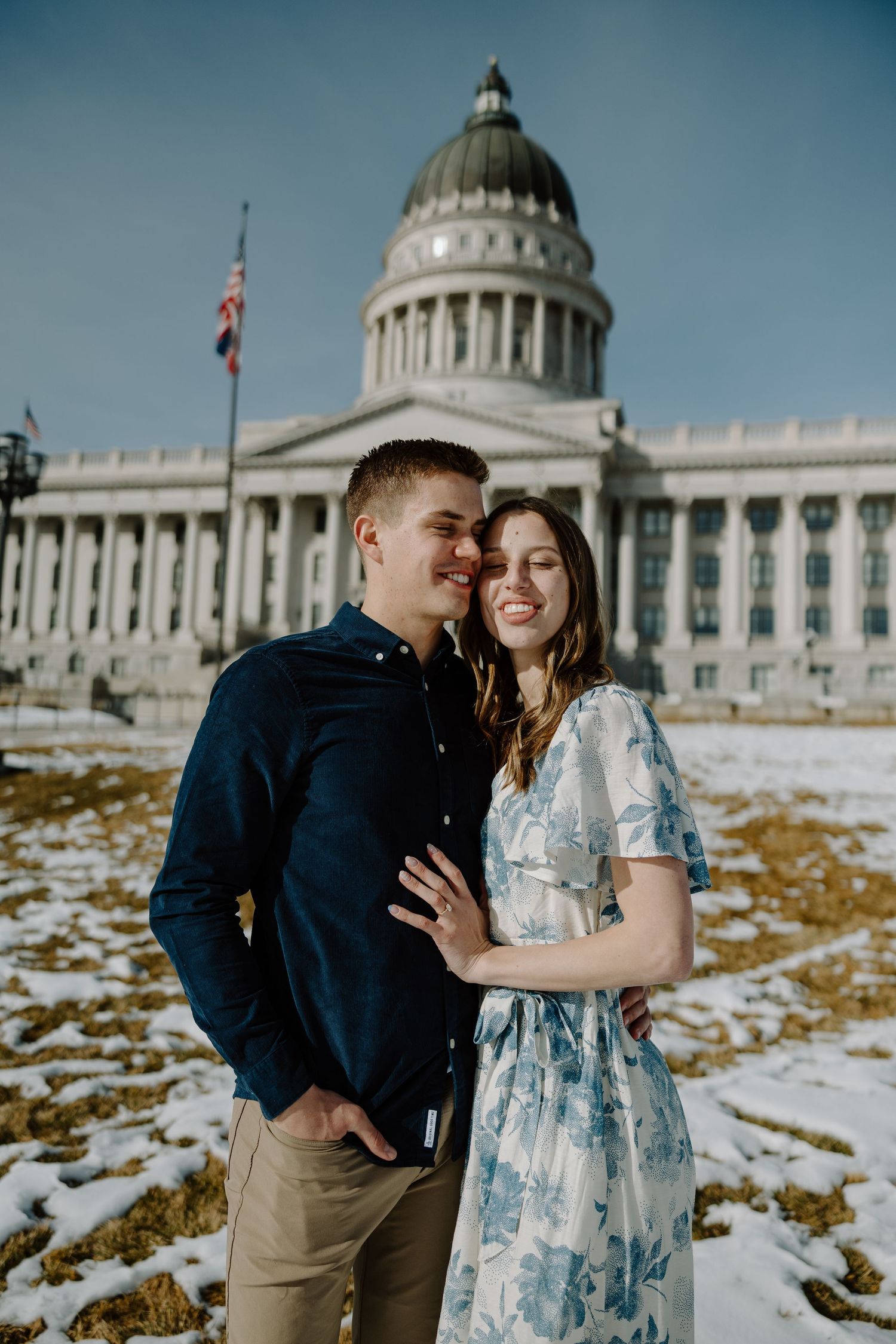 Couple embracing in front of a snow-dusted capitol building dome under a bright blue winter sky.