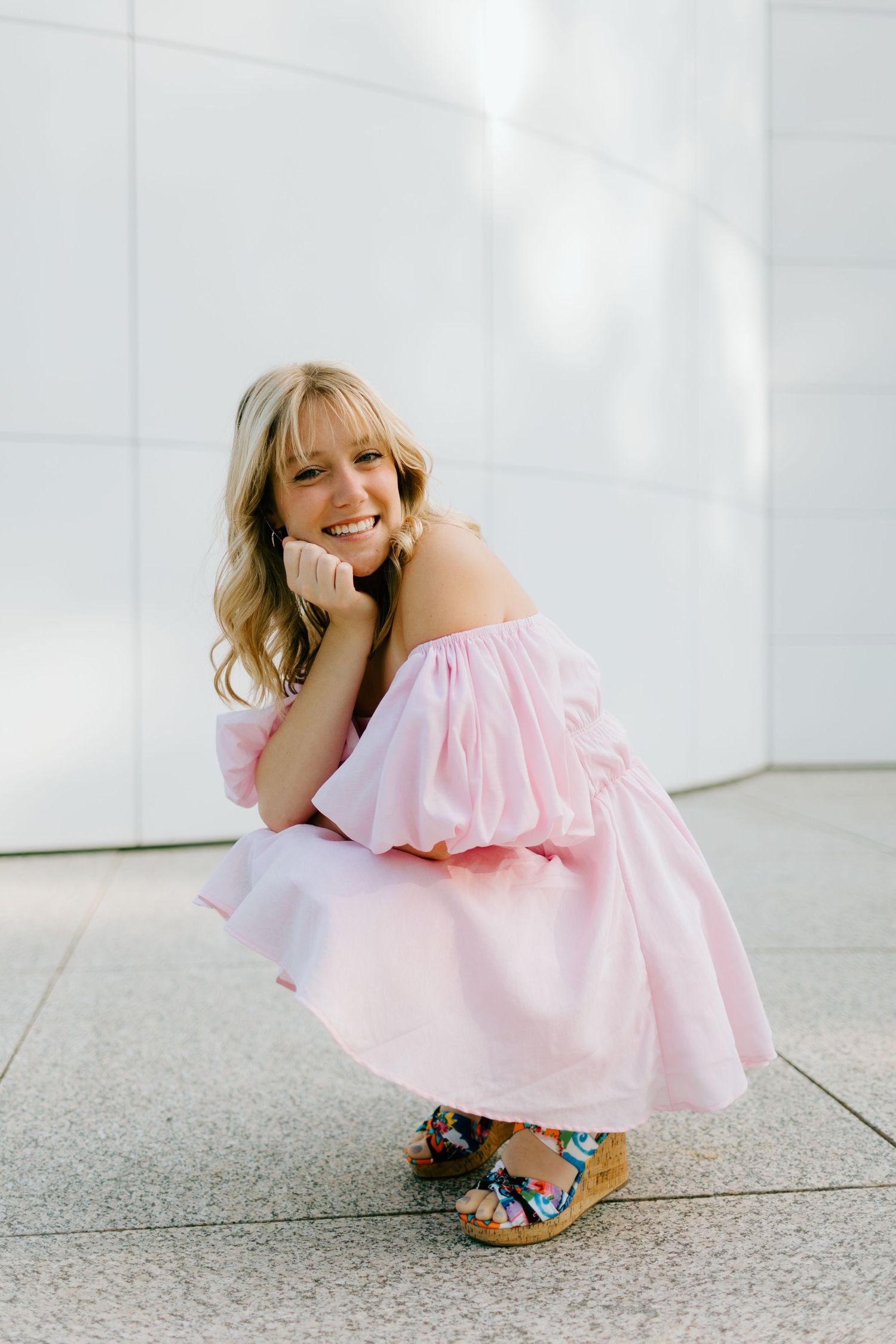Smiling blonde woman in pink off-shoulder dress crouching outdoors, wearing floral wedge sandals against a white wall.