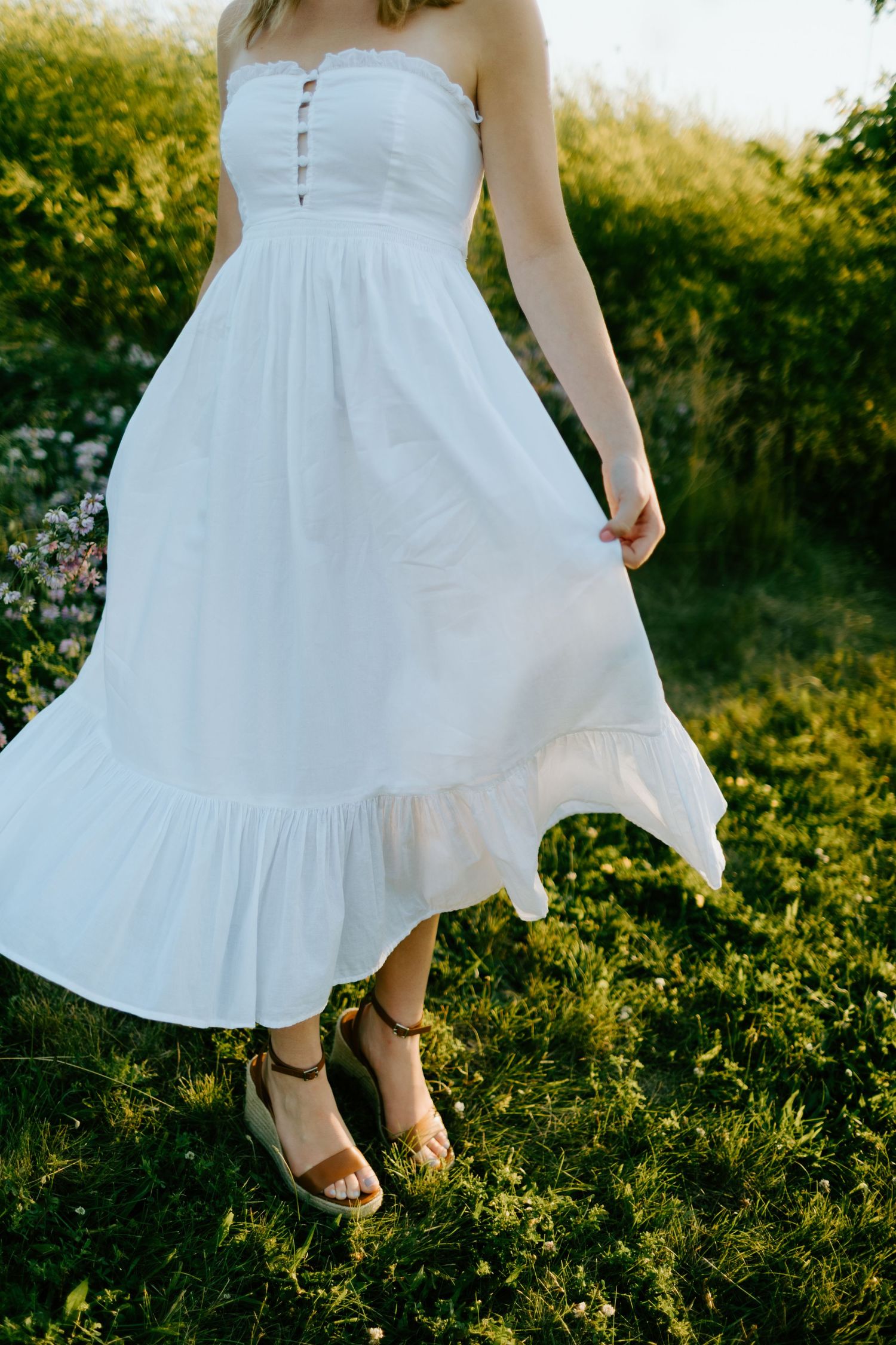 Woman in a white strapless midi dress with ruffle hem and strappy sandals standing on green grass.