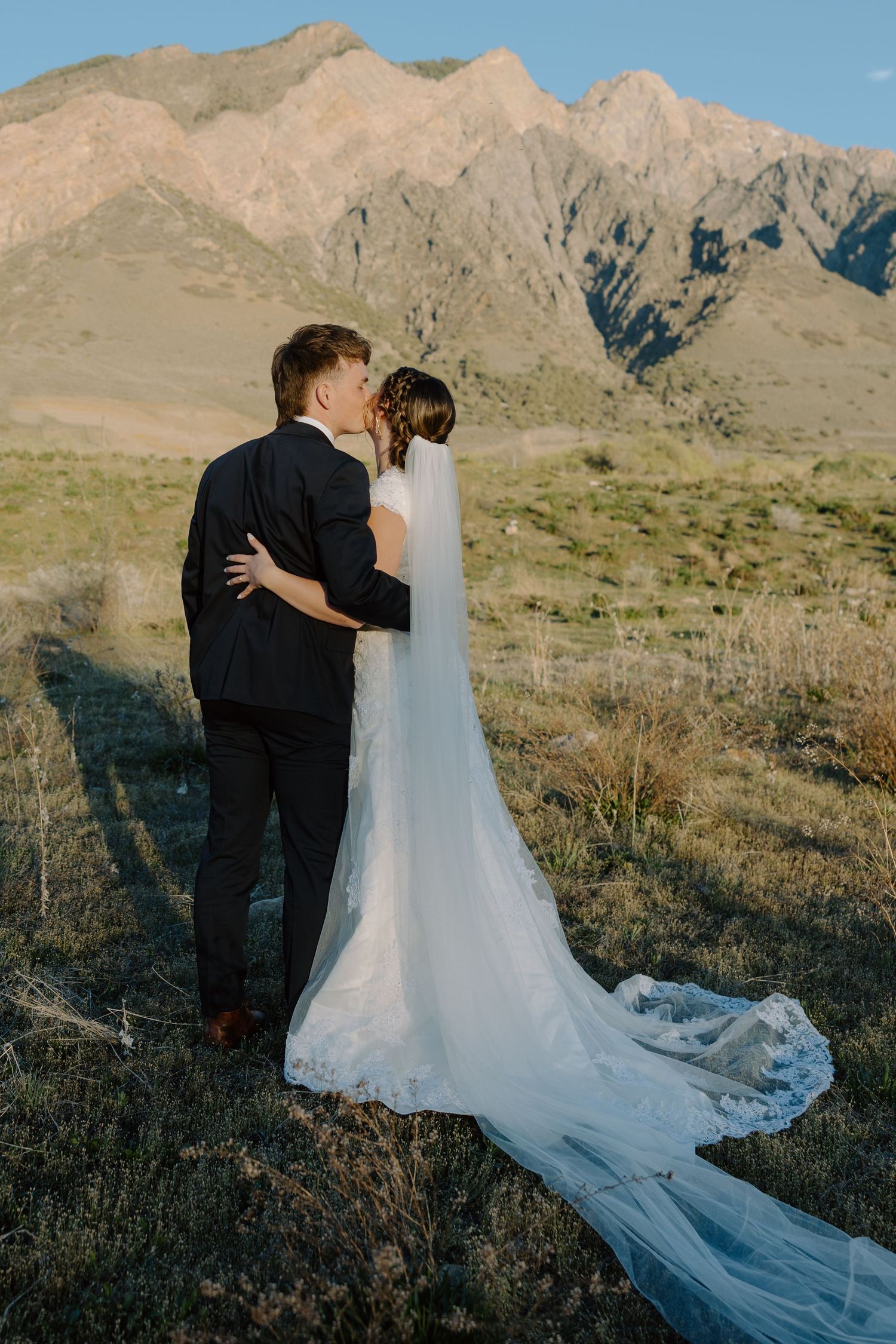 Bride and groom share a kiss with long veil trailing across a mountain meadow.