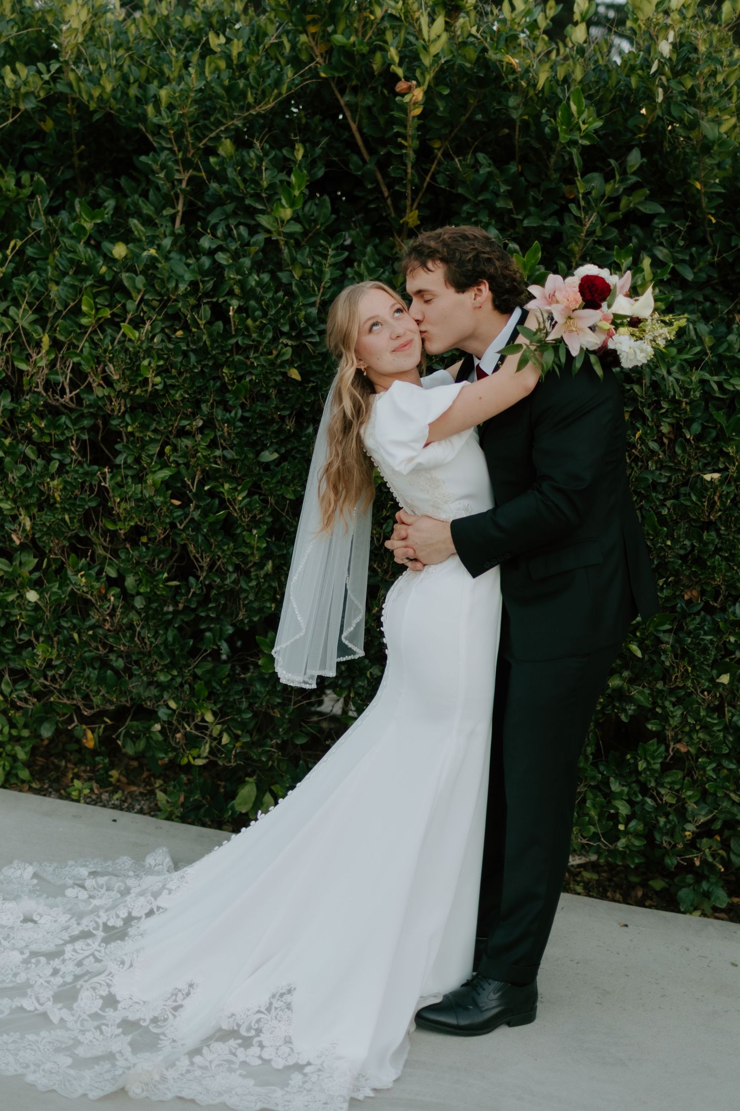 Bride in white mermaid gown with lace train and veil embraces groom in black suit kissing her cheek outdoors.