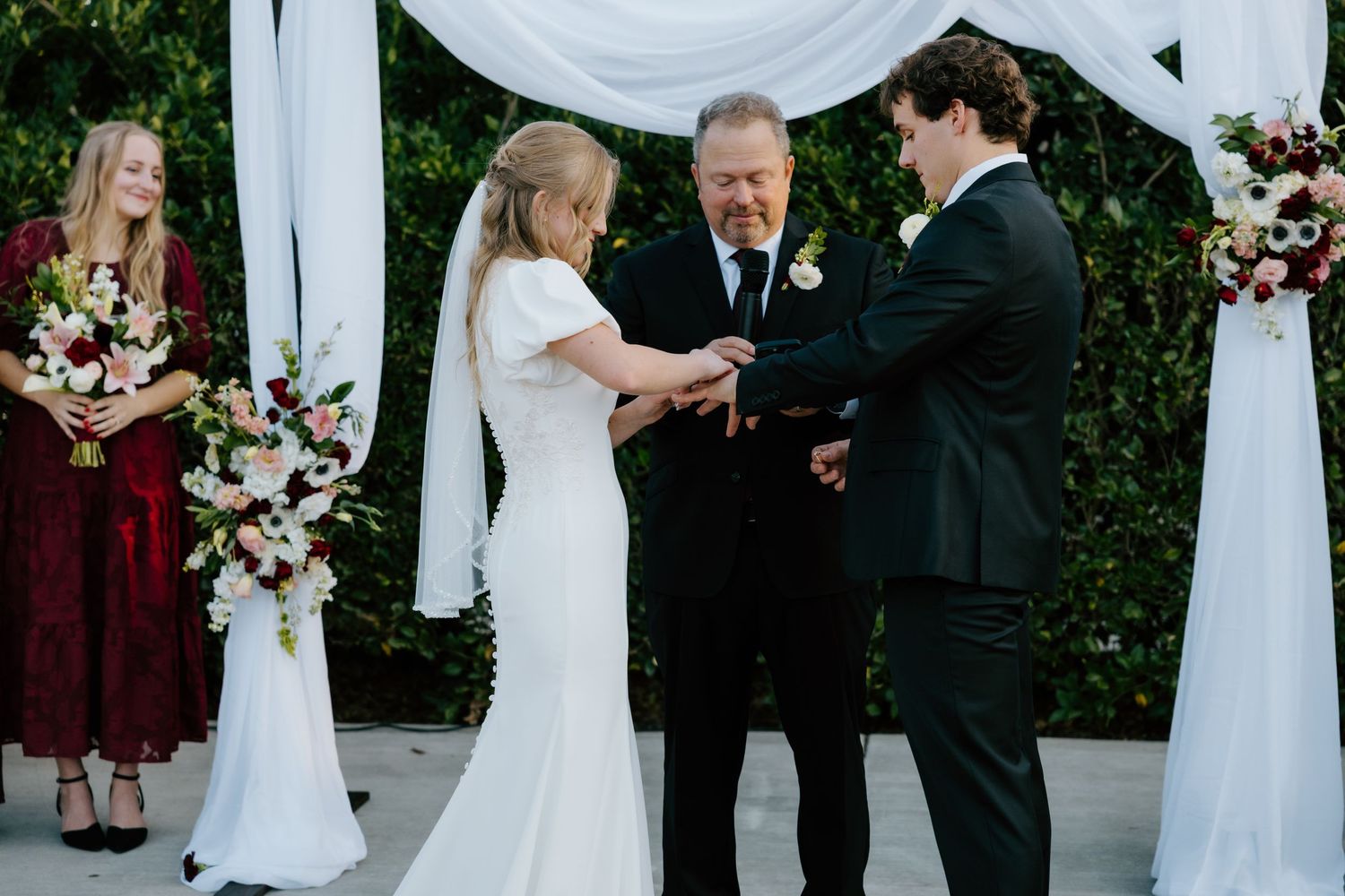 Bride and groom exchanging rings at outdoor wedding ceremony with white floral arch, officiant and bridesmaid in burgundy nearby.