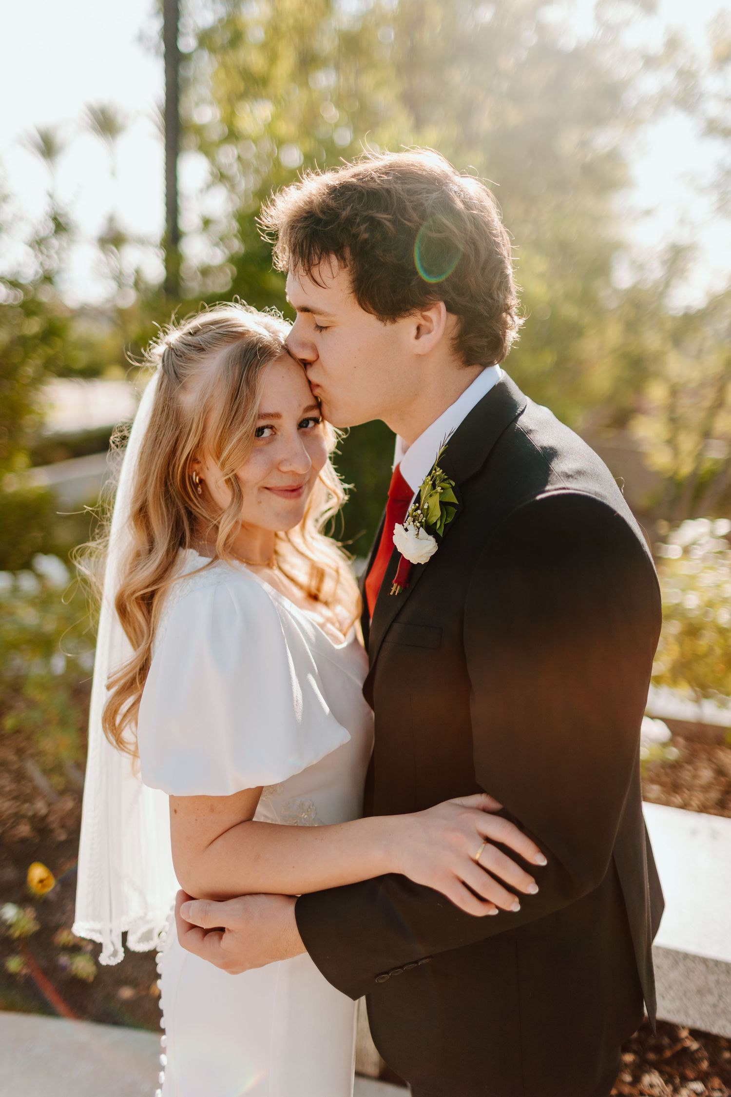 A groom in a black suit kisses his bride in a white dress and veil outdoors on their wedding day.