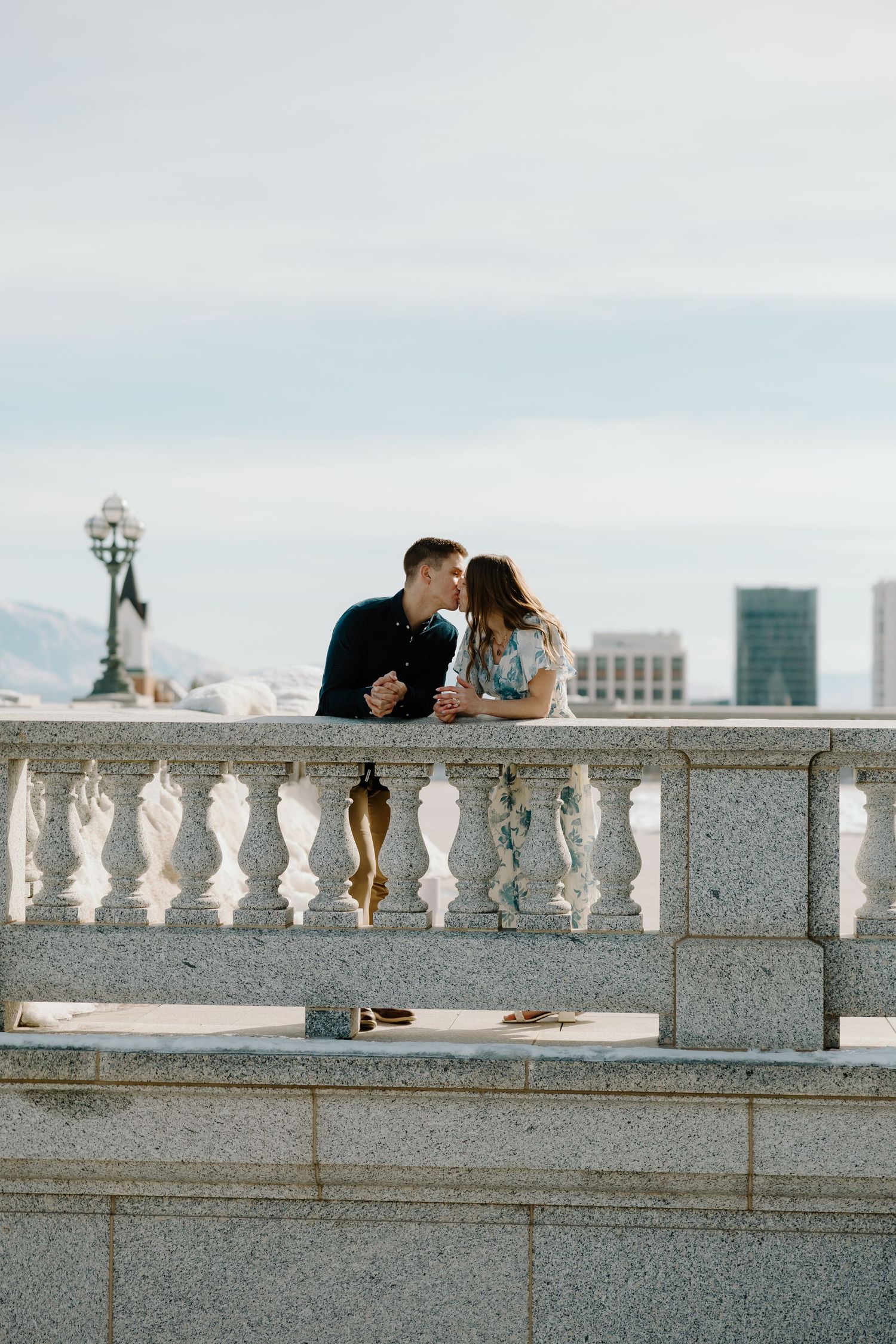 Romantic couple sharing a kiss on a stone balcony with city skyline in the background.