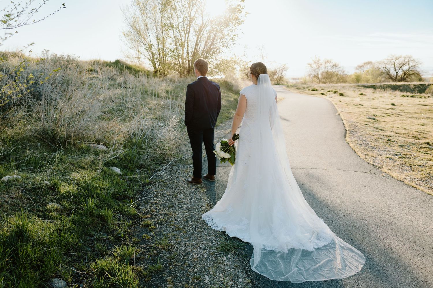 Bride and groom walking hand in hand down a sunlit path on their wedding day.
