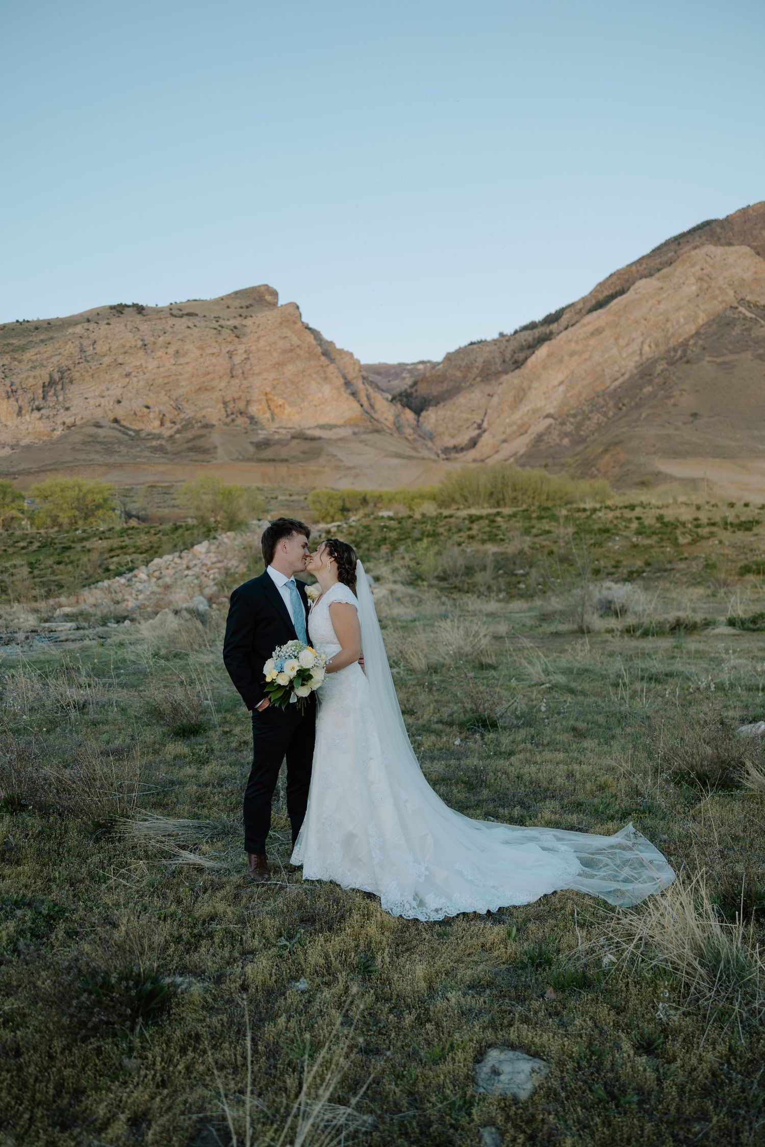 Bride and groom share a romantic moment in a scenic desert landscape with rocky mountains in the background.