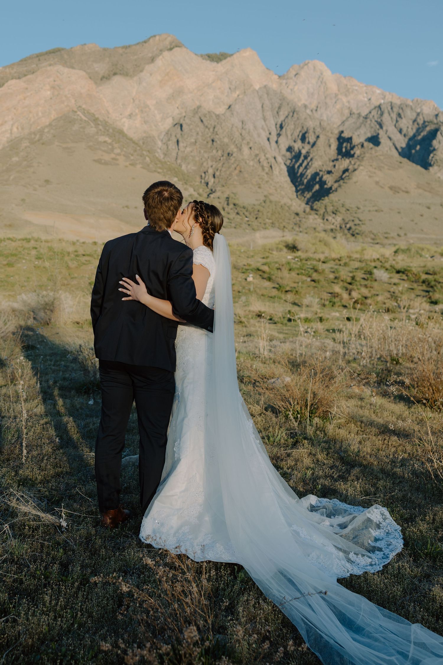 Bride and groom kissing with backs to camera in golden mountain landscape at sunset.