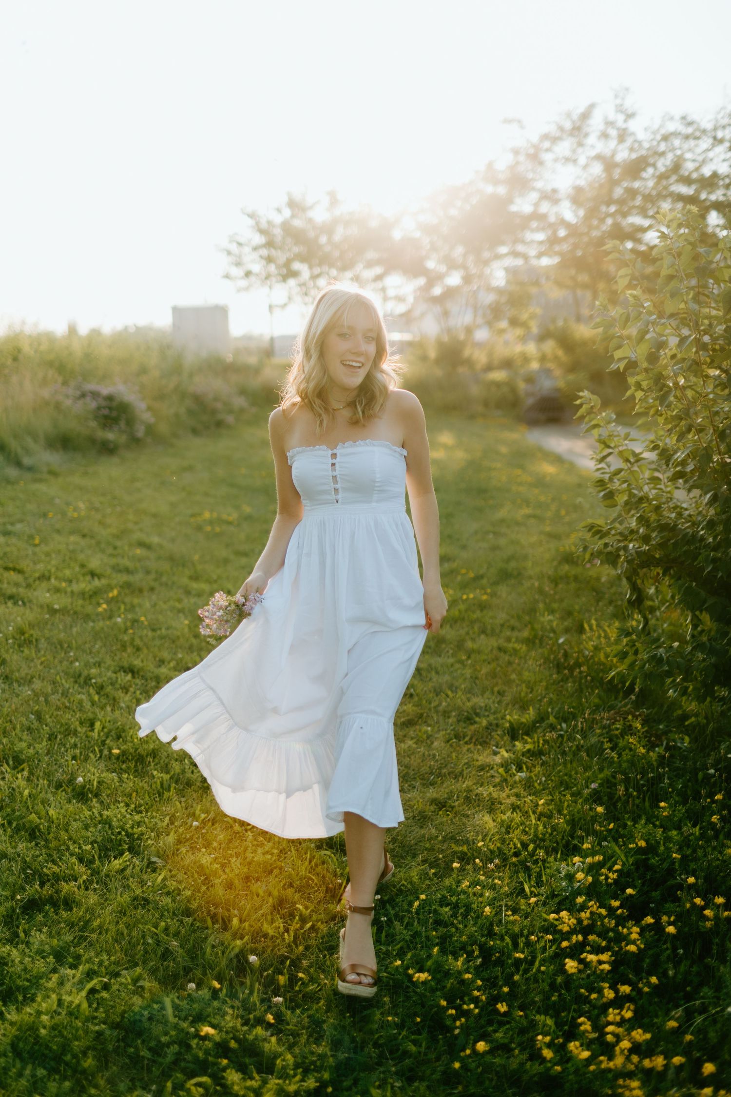 Smiling blonde woman in white strapless midi dress walking through green field at golden hour sunset.