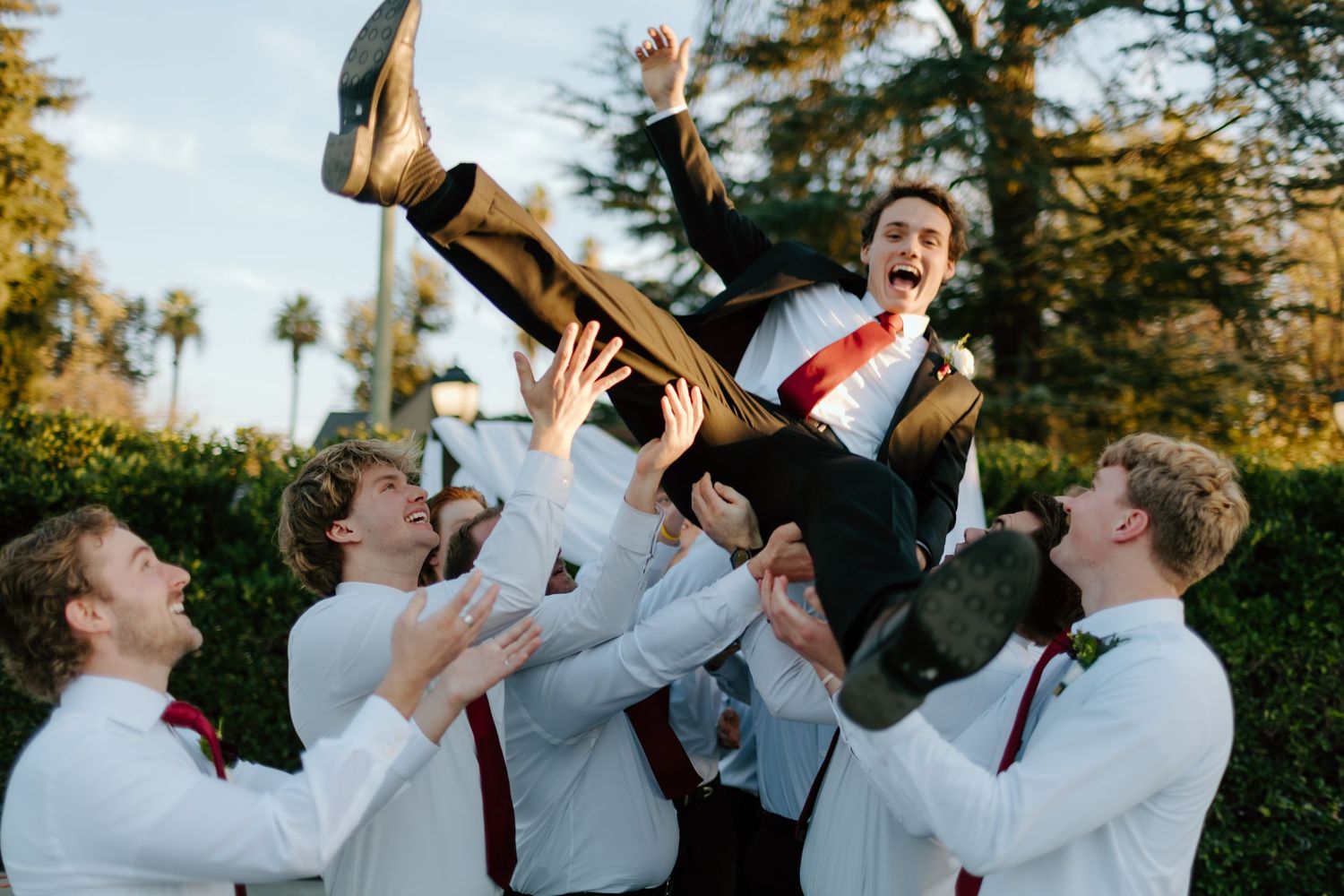 Groom lifted by groomsmen in white shirts and red ties outdoors, celebrating wedding day with joy.