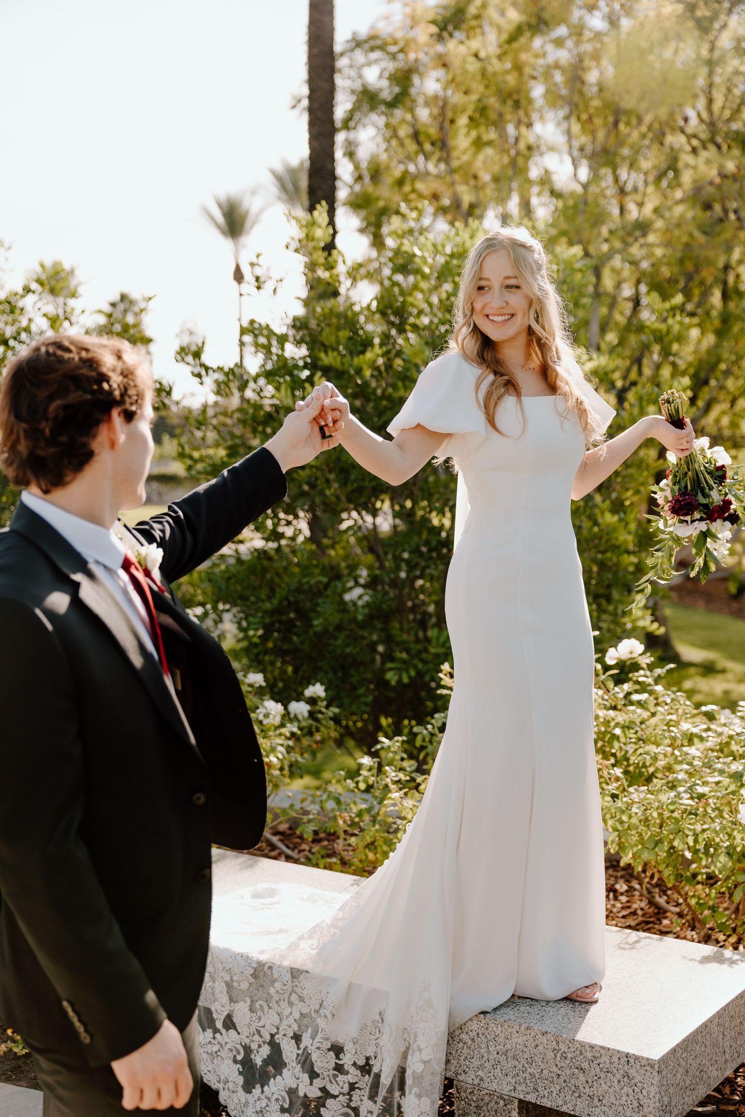 Bride in white gown stands on stone ledge holding groom's hand and bouquet in sunlit garden.