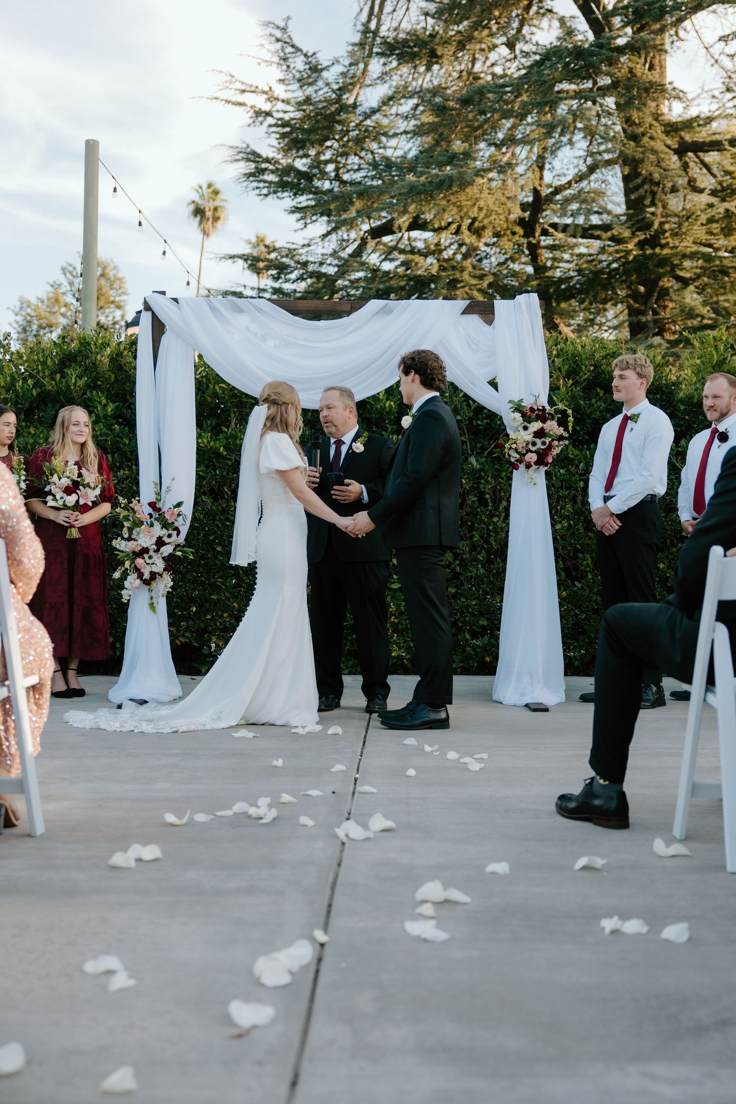 Bride and groom exchange vows at an outdoor ceremony beneath a white draped arch with rose petal aisle.