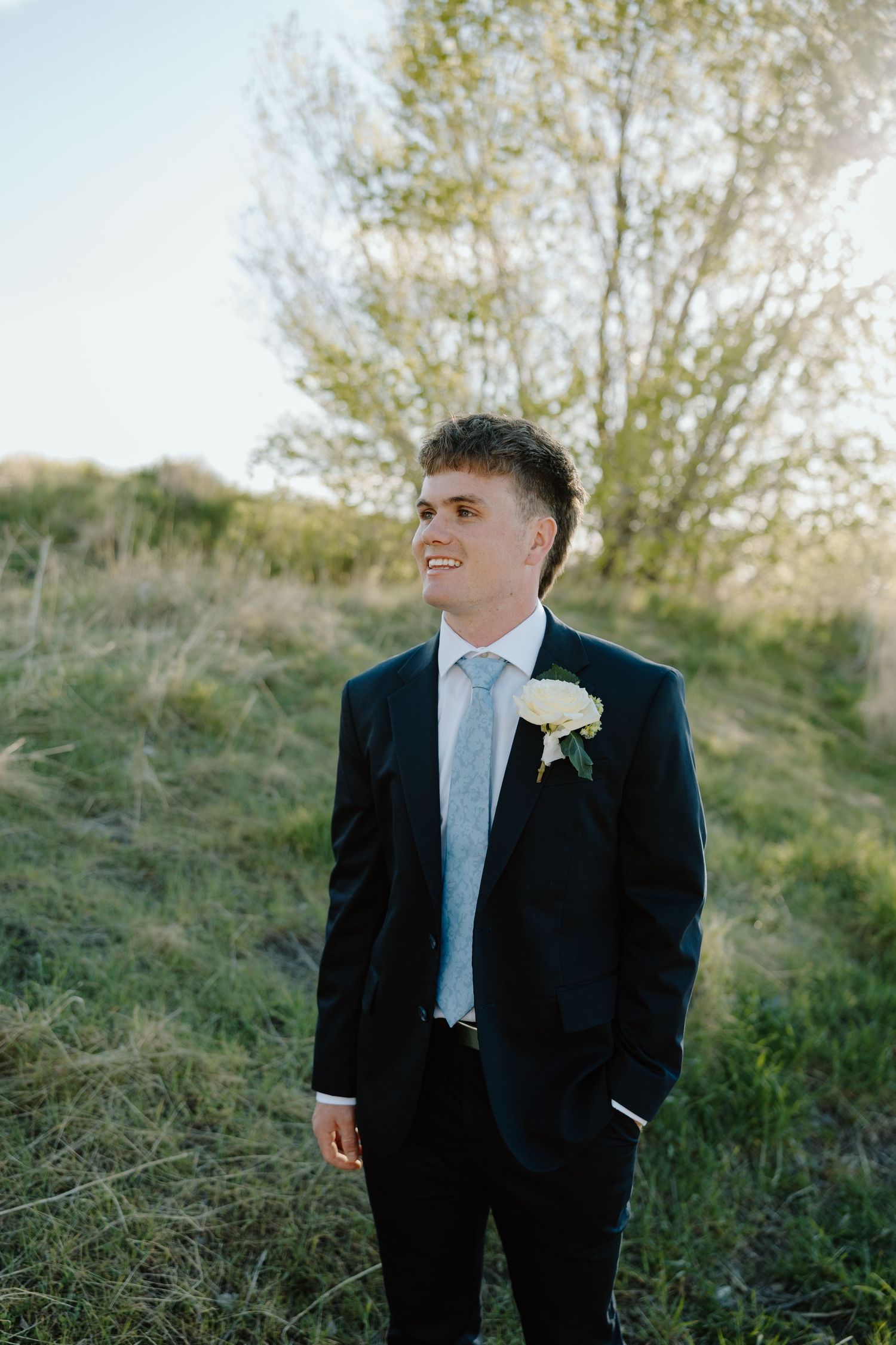 Smiling groom in black suit with white boutonniere standing in green field with trees in background.