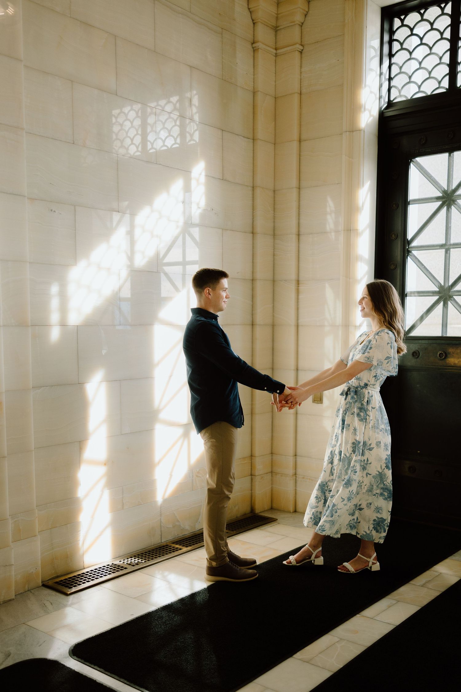 Couple holding hands in sunlit ornate building hallway, woman in blue floral midi dress, man in navy jacket.