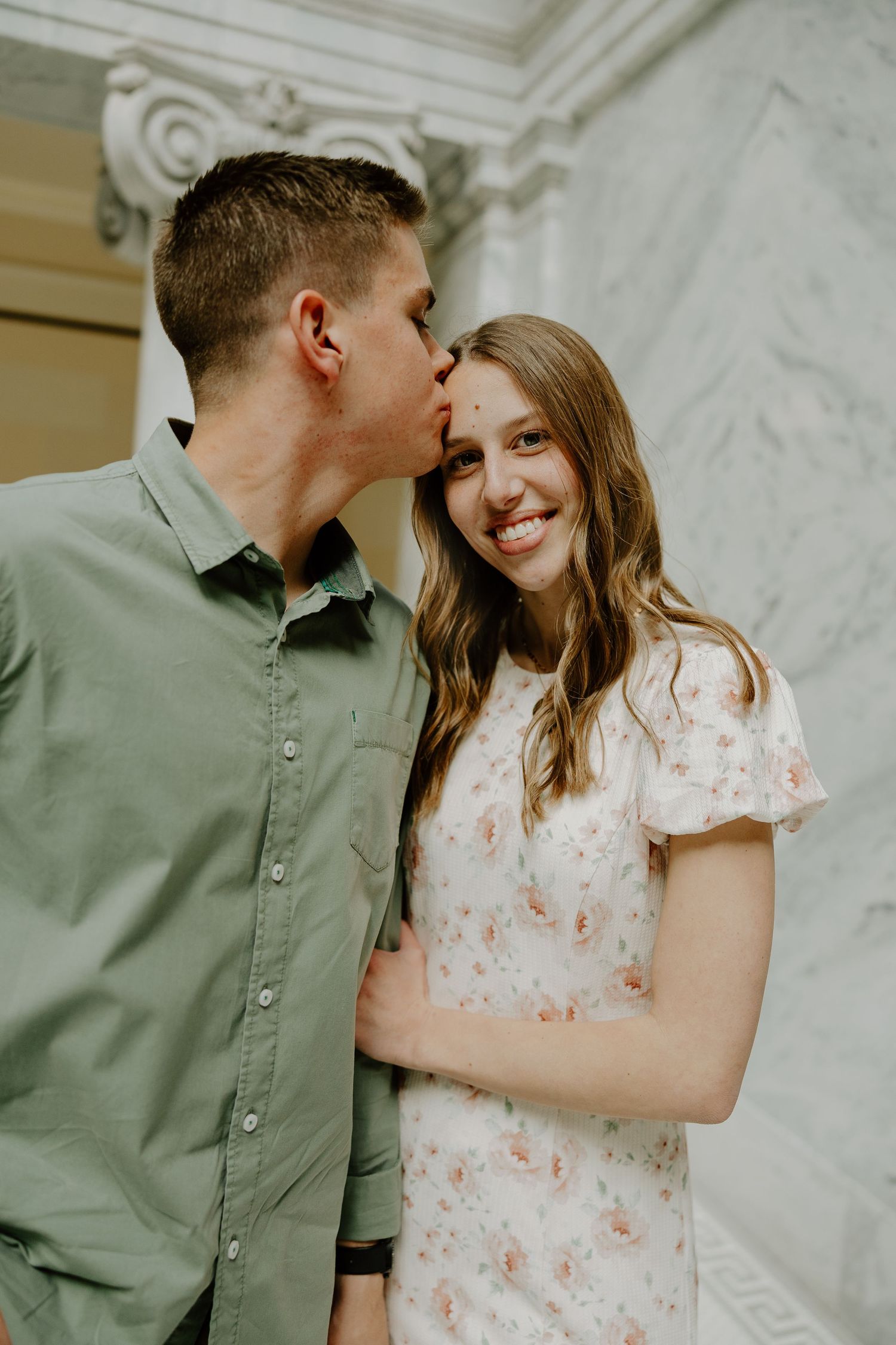 A couple shares a tender moment as a man kisses a smiling woman on the forehead near marble columns.