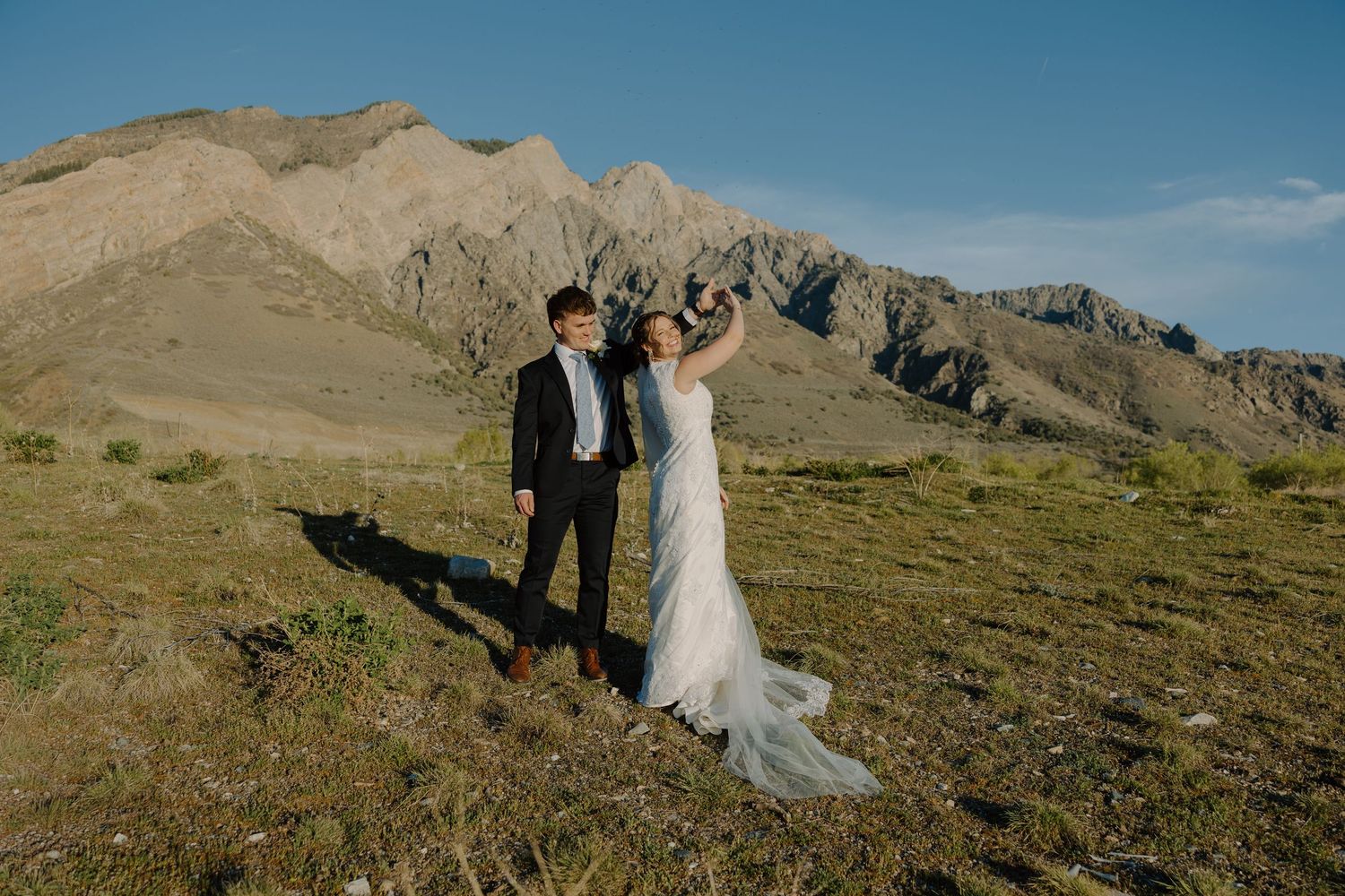 Joyful bride and groom celebrating with raised arms on a hillside with mountain views.