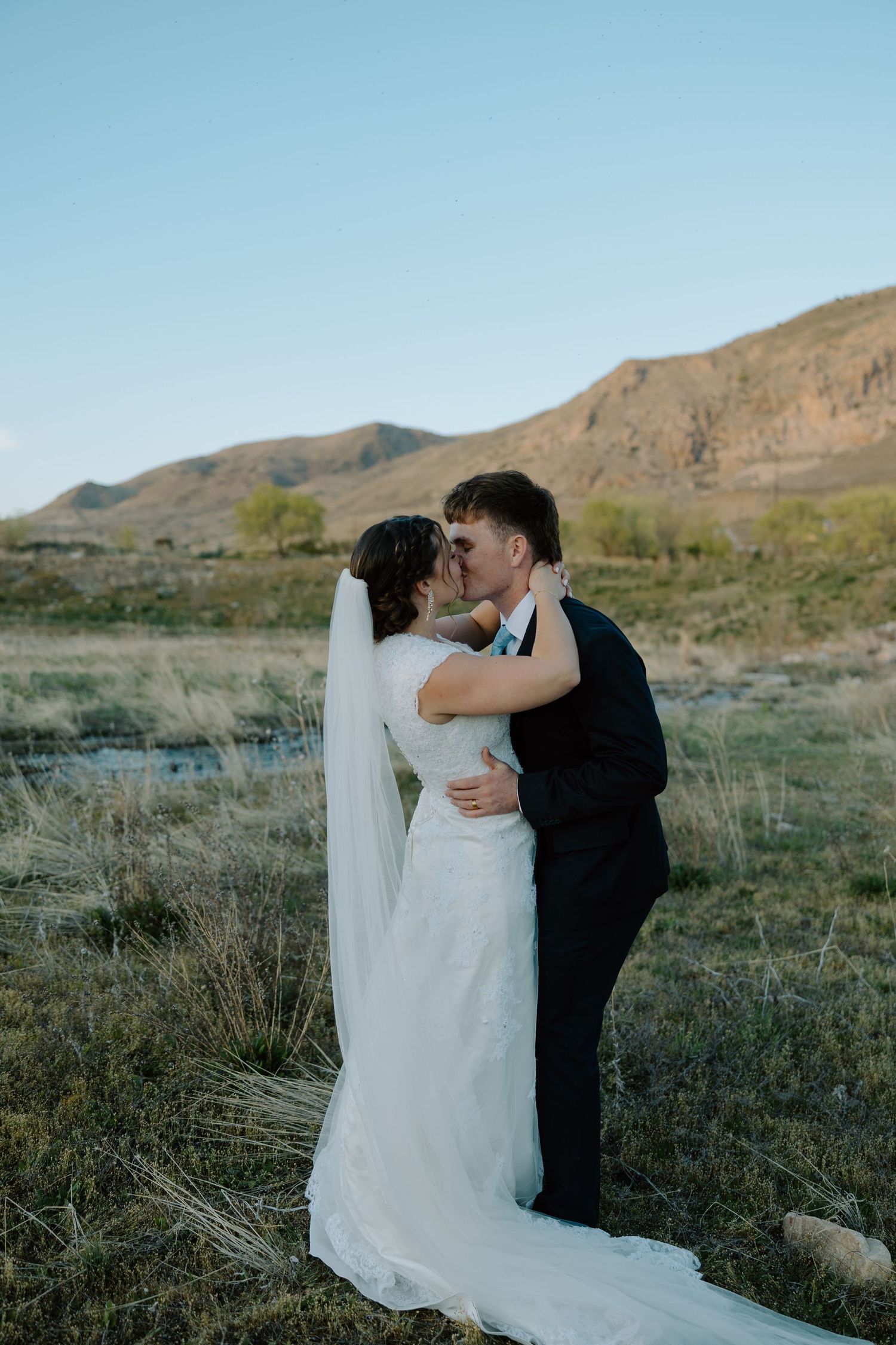 Newlyweds share a romantic first kiss in a rustic field with sunlit mountains in the background.