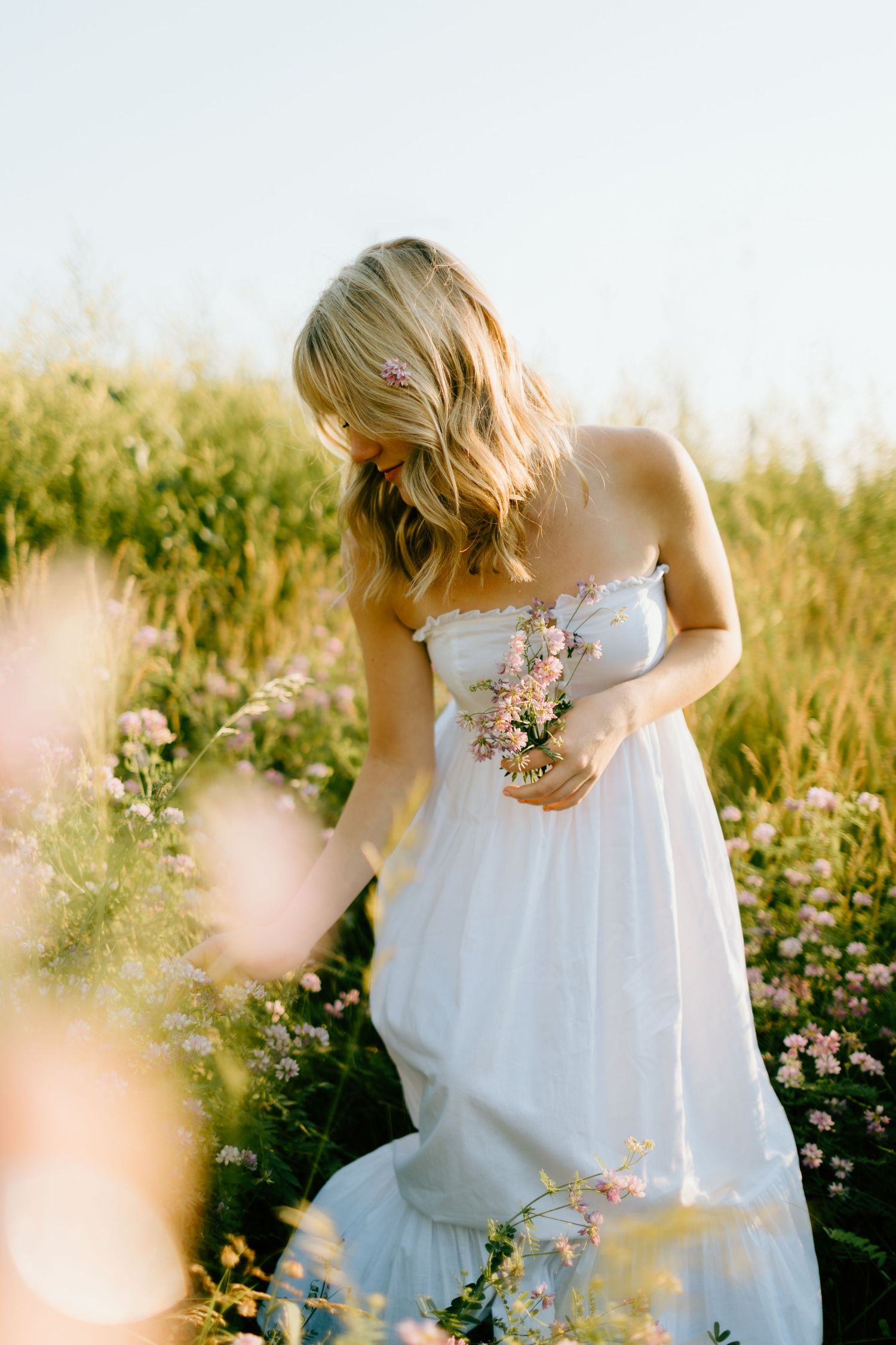 Blonde woman in white strapless gown holding wildflowers in a sunlit meadow field.