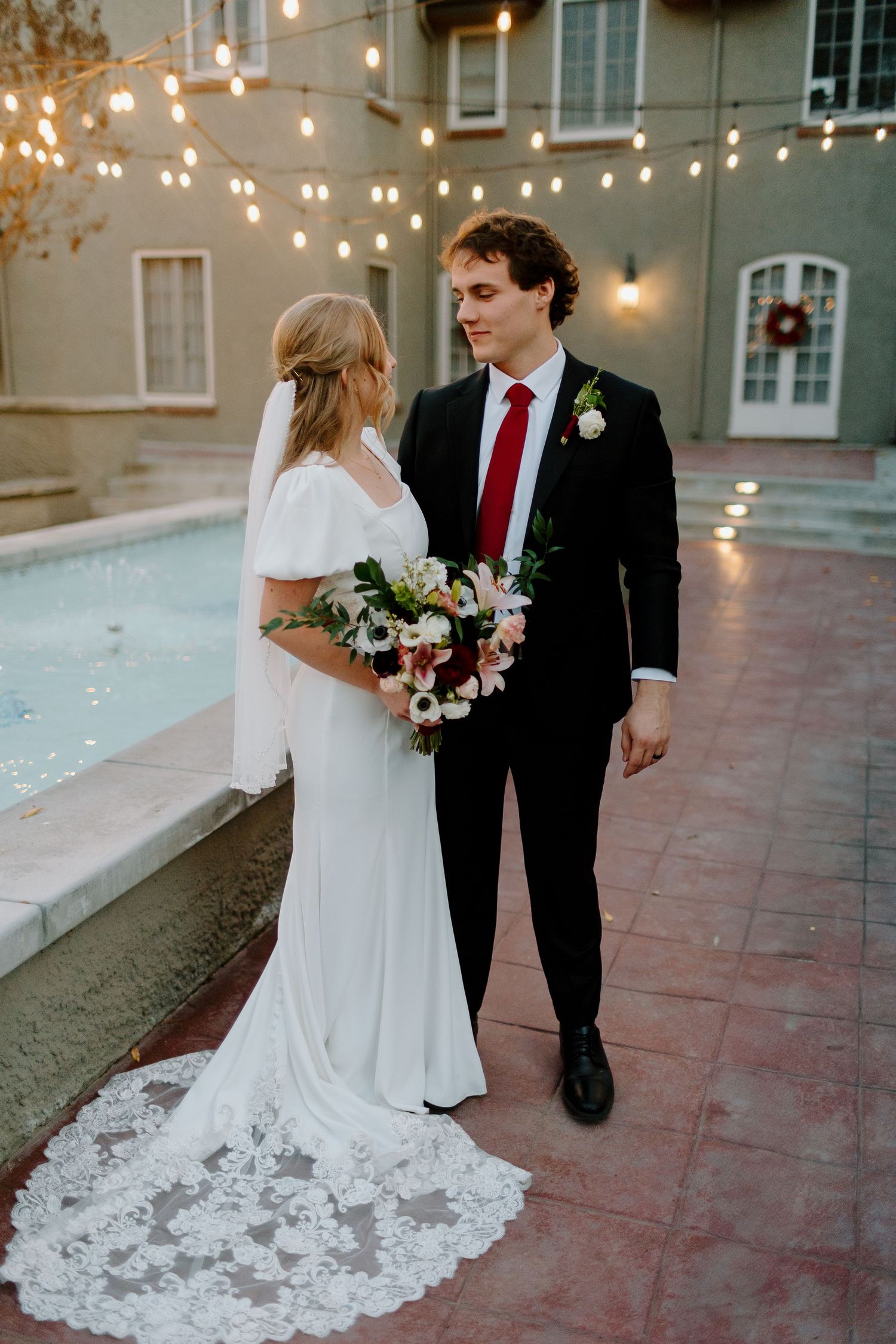 Bride and groom walk hand-in-hand near a fountain at dusk, bride holding a white and pink bouquet.