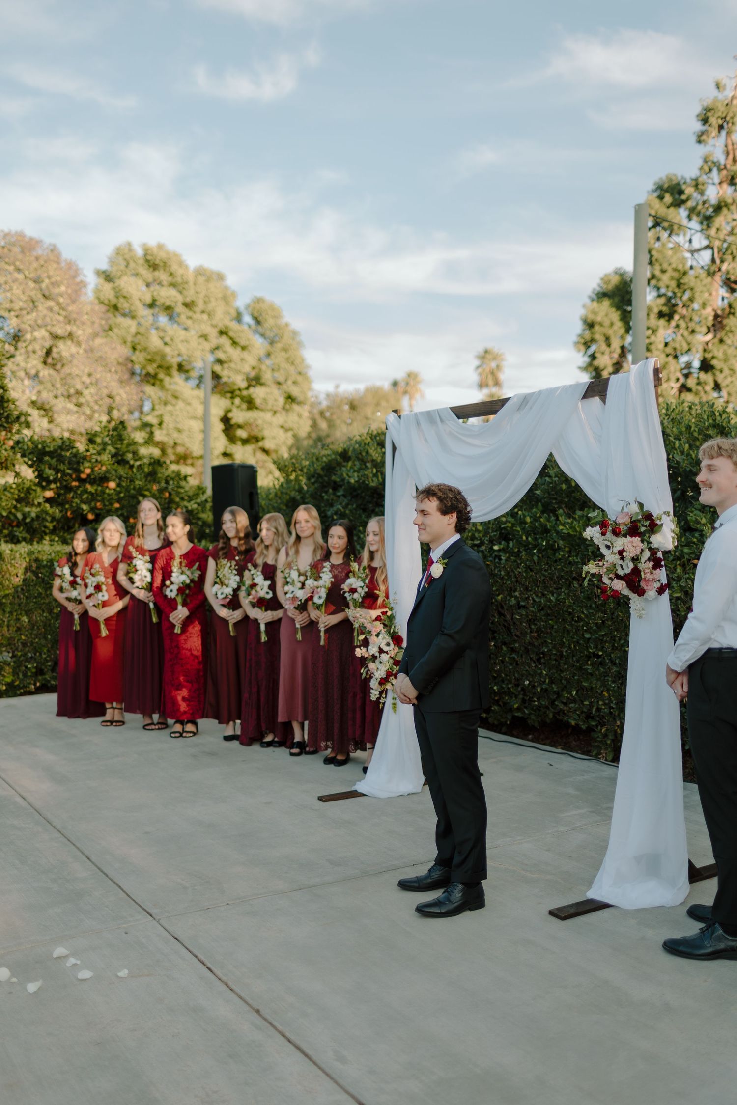 Outdoor wedding ceremony with bridesmaids in red dresses standing beside a white draped arch with floral arrangements.