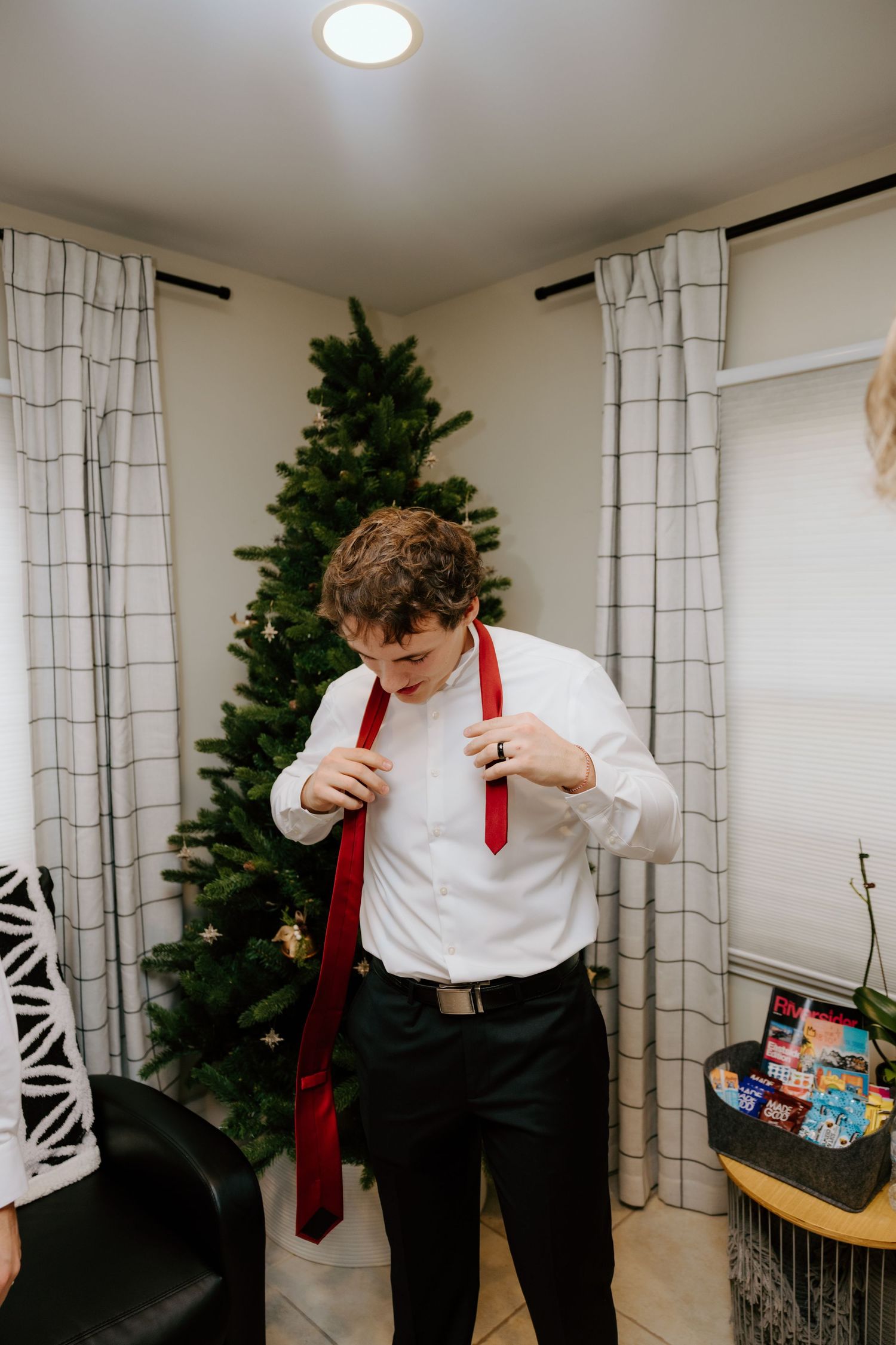 Young man in white shirt adjusting red tie near Christmas tree in decorated living room.