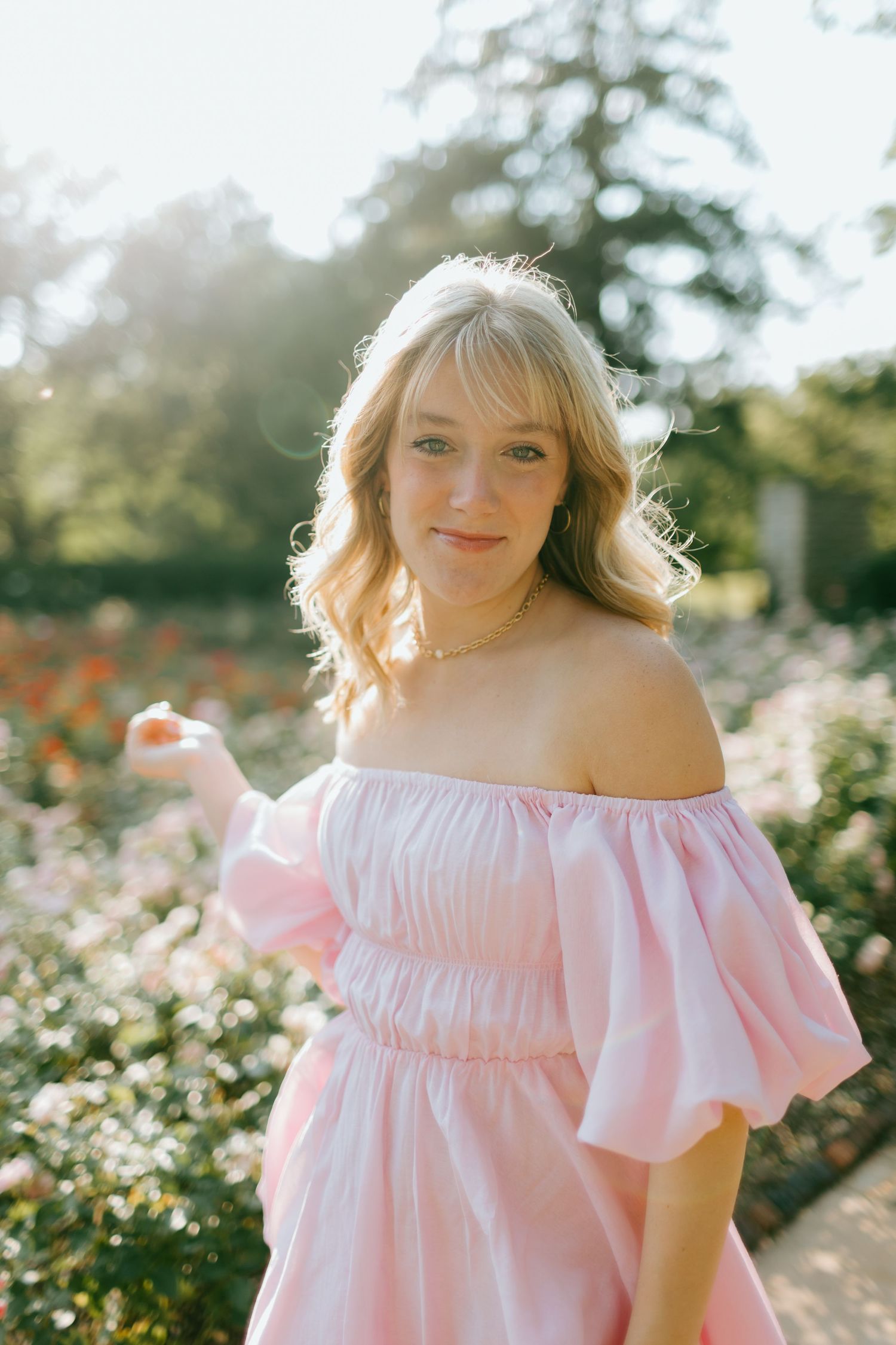 Young blonde woman in pink off-shoulder dress posing in a sunlit garden with blooming flowers.