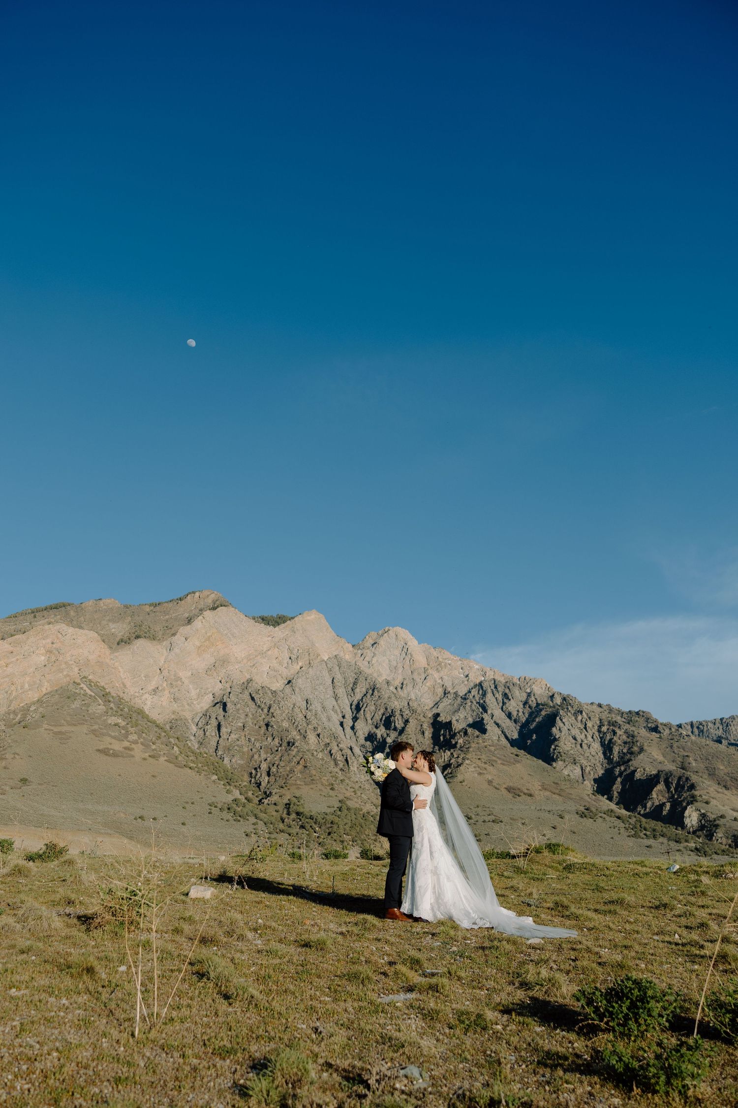 Wedding couple embracing with dramatic rocky mountain backdrop under a clear blue sky.