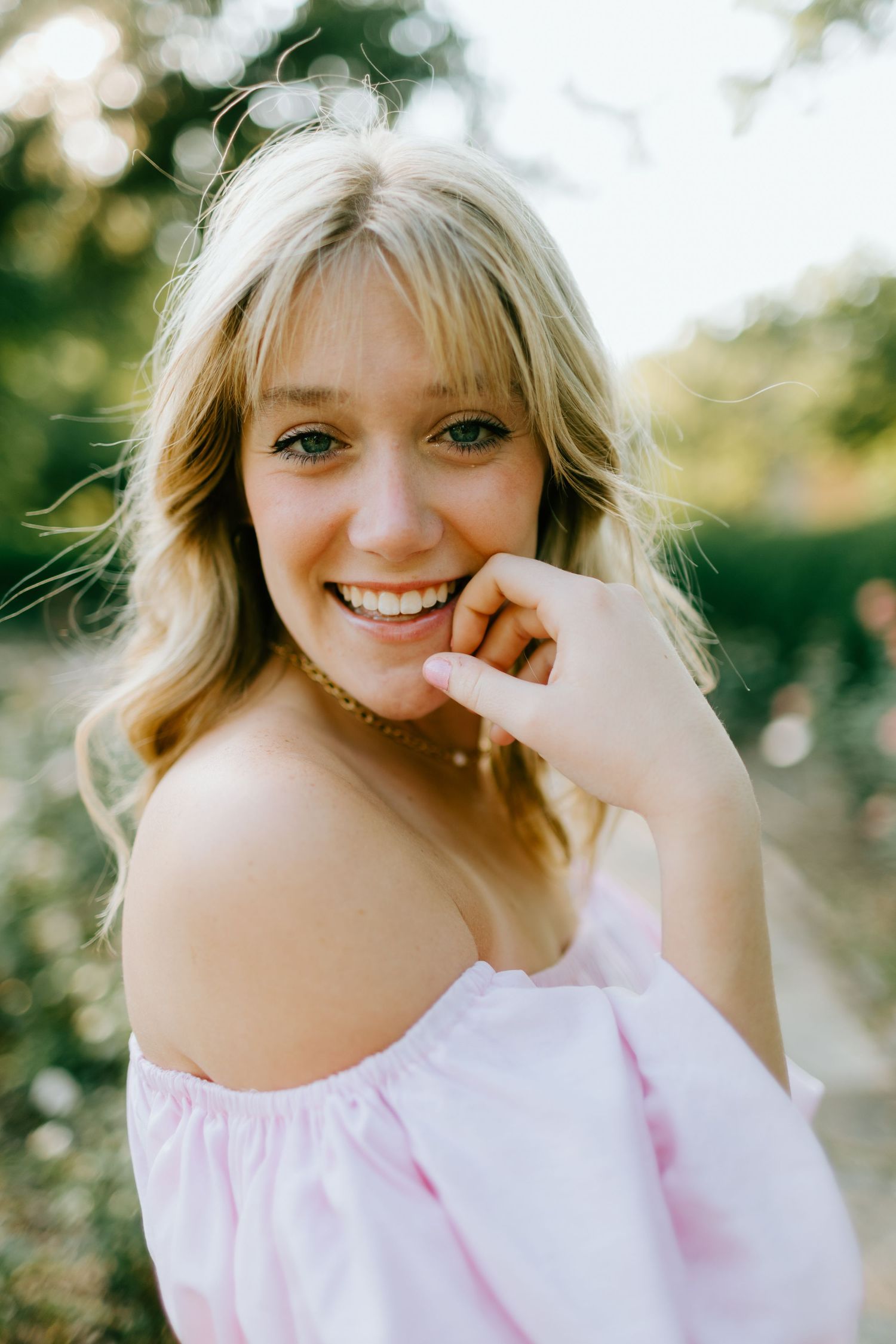 Smiling young woman with blonde hair wearing an off-shoulder pink top, posing outdoors in soft natural light.