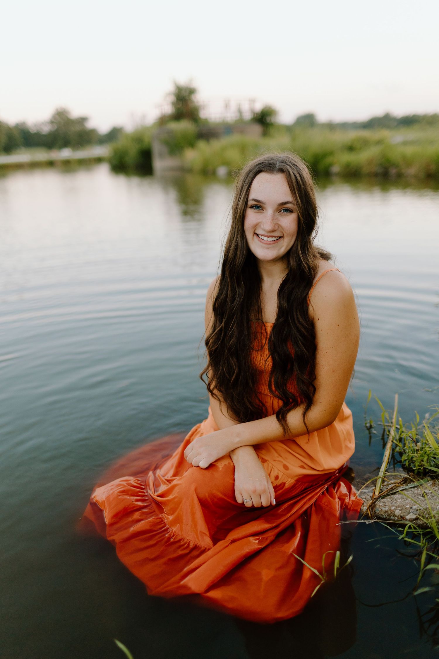 Young woman in orange dress sitting by a calm lake shore, smiling with long dark hair.