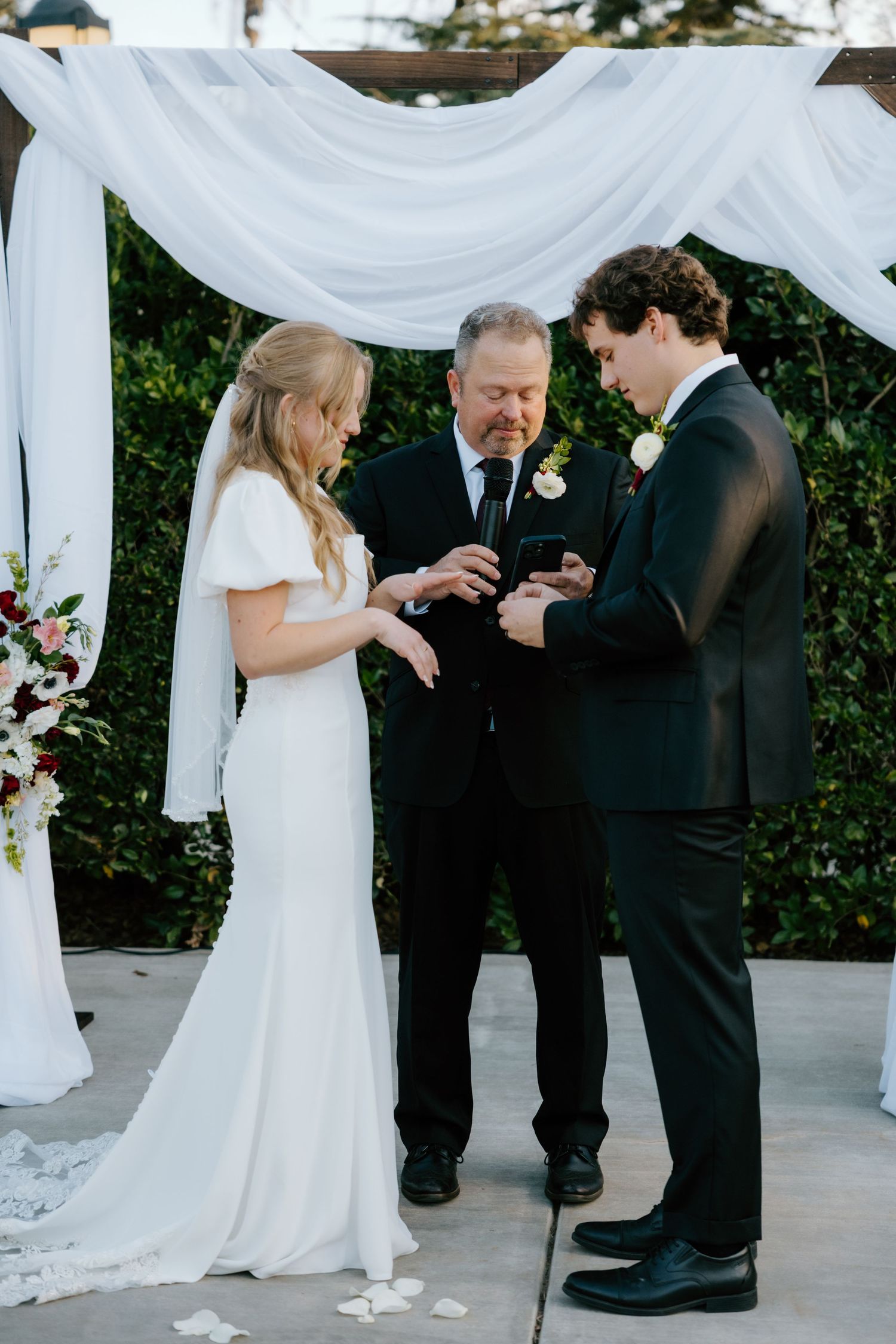 Officiant places wedding ring on bride's finger as couple exchanges vows under a white draped ceremony arch.