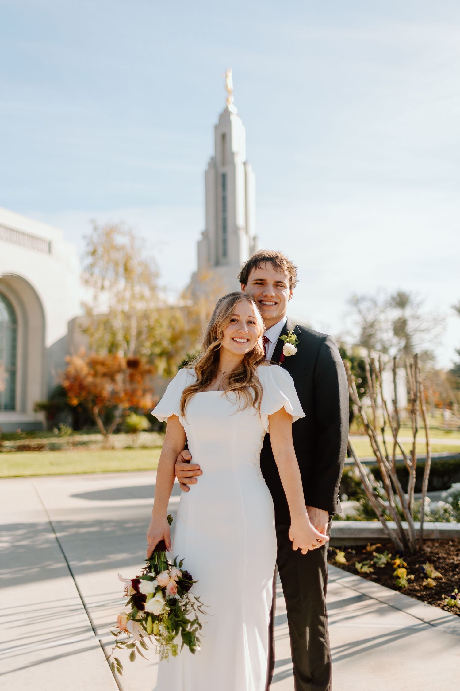 Bride and groom holding hands and smiling in front of an LDS temple on their wedding day, bride holding a floral bouquet.