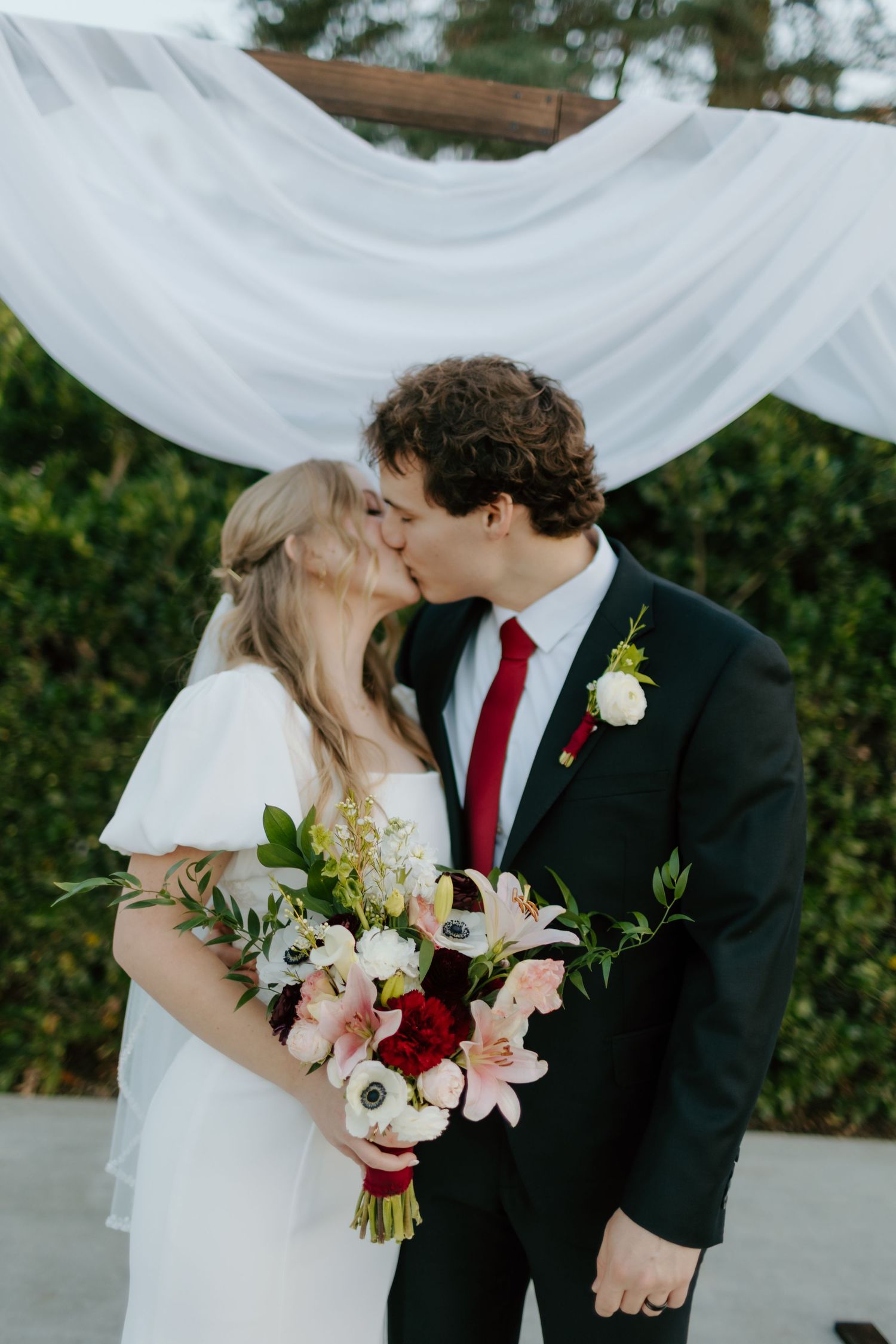 Bride and groom sharing a kiss under white draped arch, bride holding colorful bouquet with anemones.