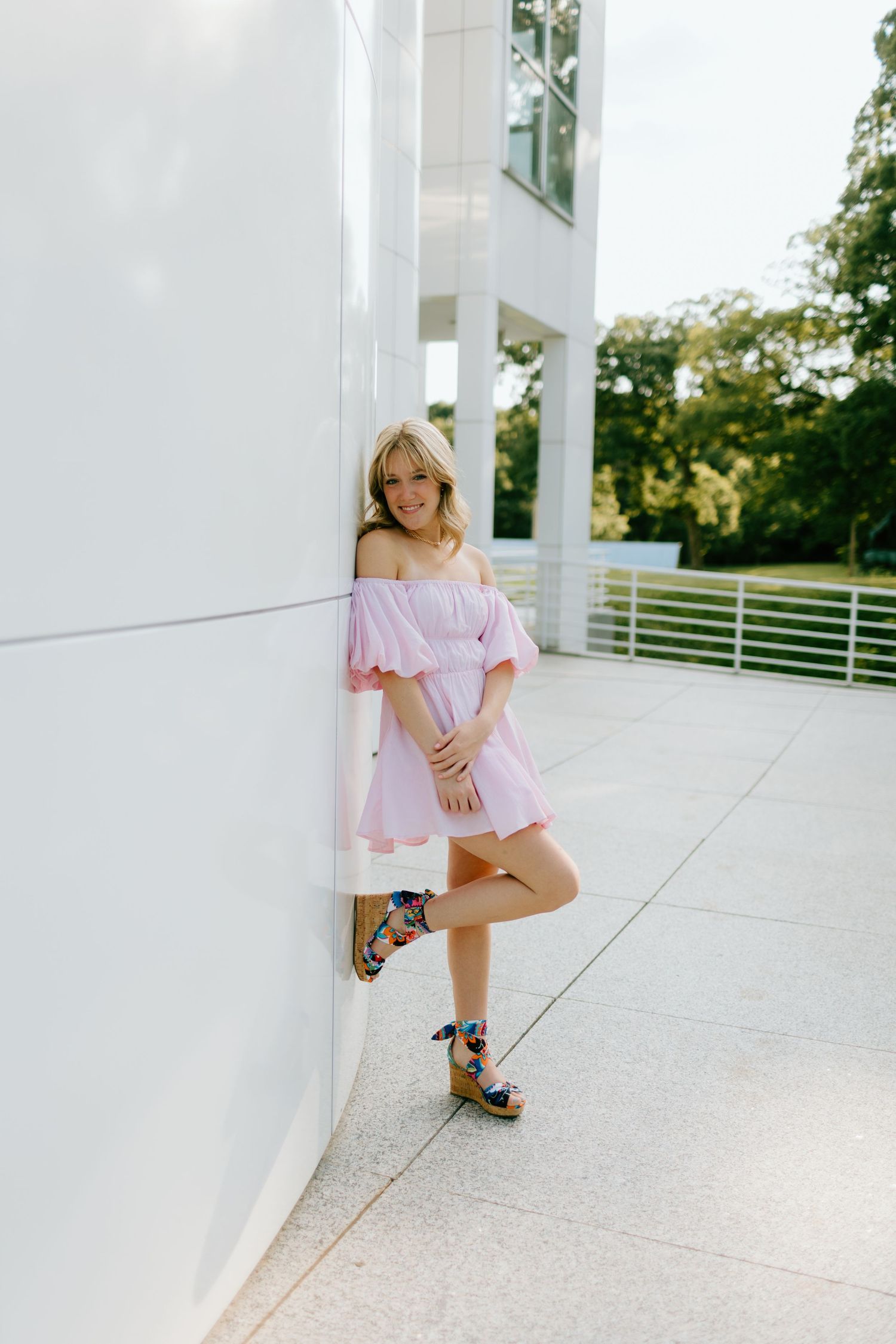 Blonde woman in pink off-shoulder mini dress and colorful platform sandals leaning against white wall outdoors.